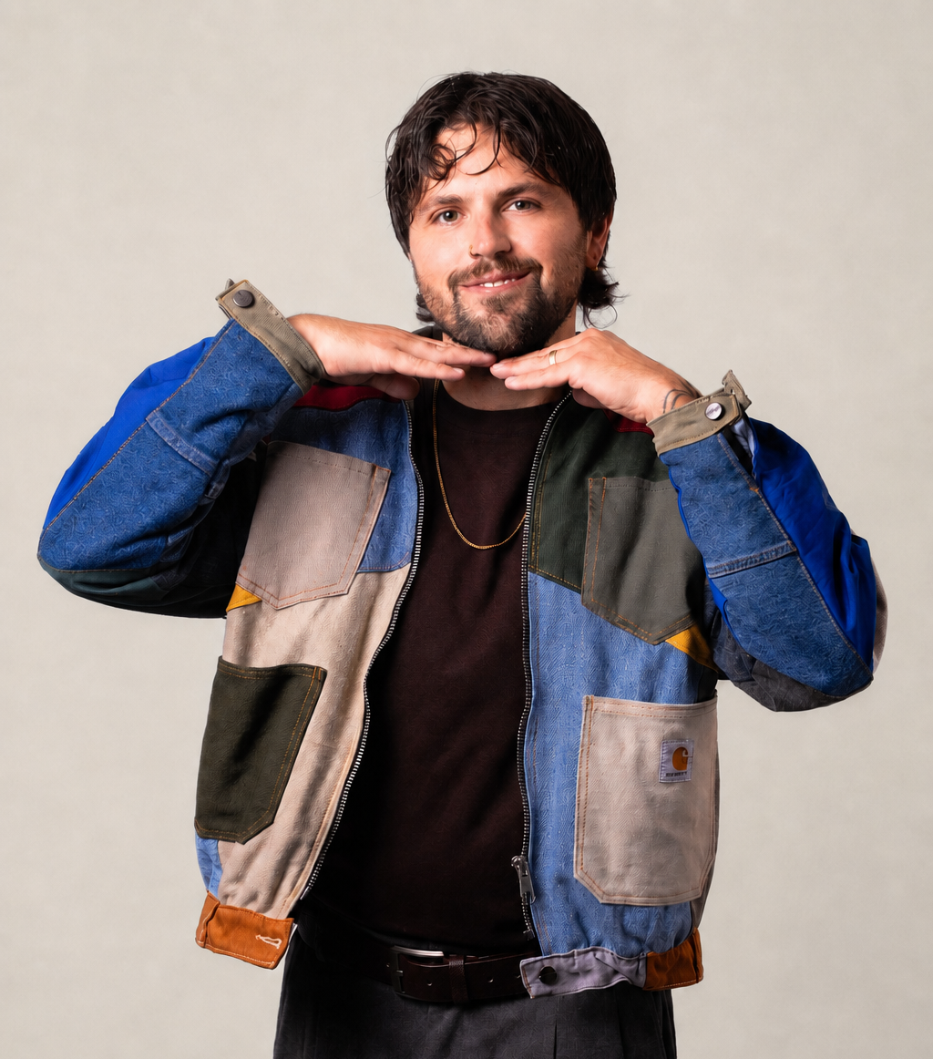 A man with dark hair and a beard smiling, making a pose with his hands under his chin, wearing a multicolored patchwork jacket and a black shirt against a plain background.