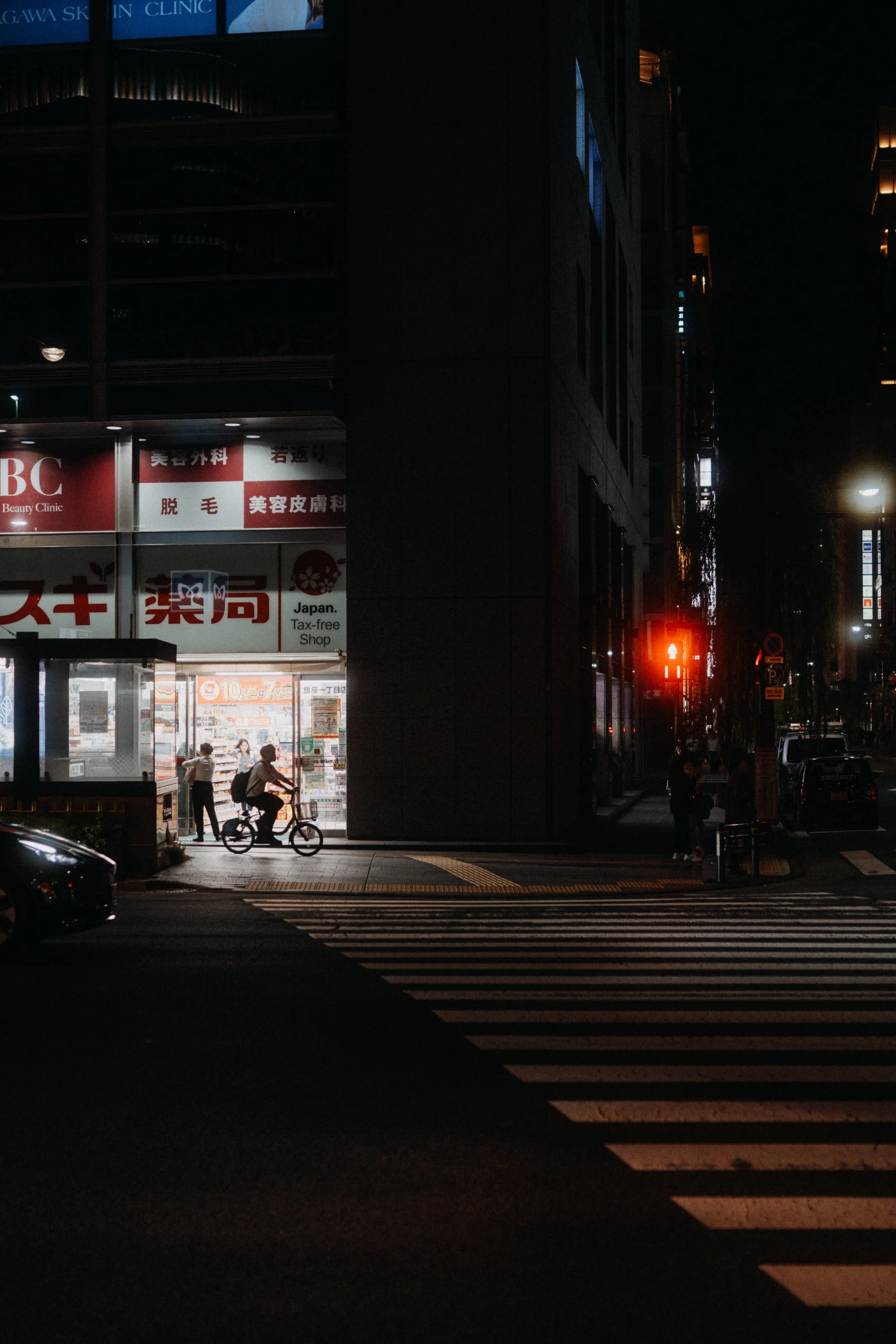 Nighttime city scene showing a crosswalk, a bicycle rider, pedestrians, illuminated shop windows, and a traffic signal with a red hand symbol.