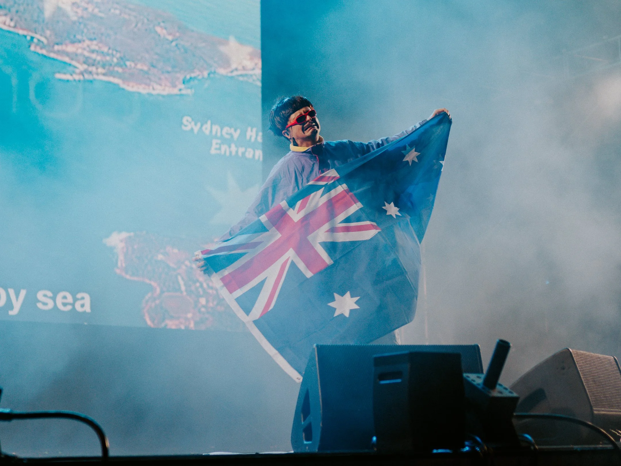 A performer on stage holding an Australian flag, wearing red sunglasses, and making a sad face. There is a large screen in the background with part of a map and the text 'Sydney H...'. The scene is filled with fog or smoke.