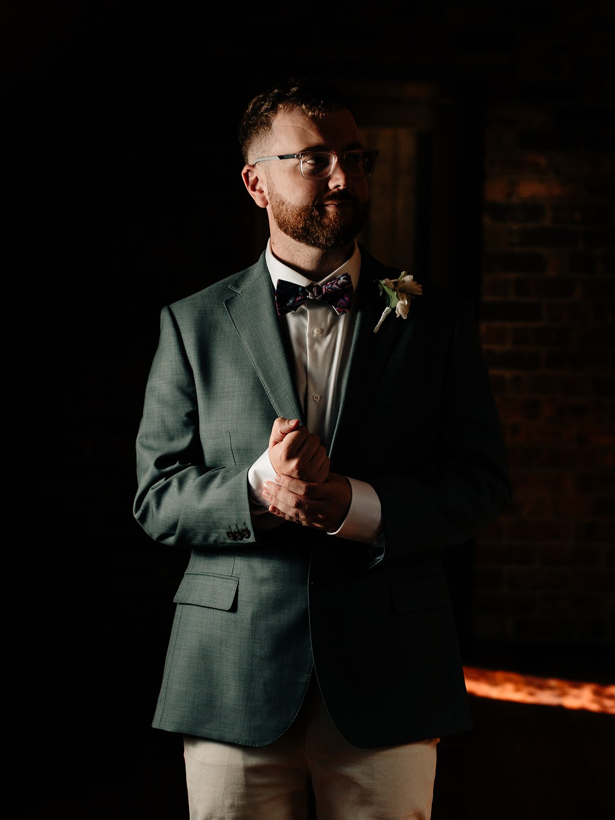 A man in a gray suit, white shirt, and bow tie stands in a dimly lit room with a dark brick wall background. He has glasses and a boutonniere, and is holding his lapel with one hand, looking to the side.