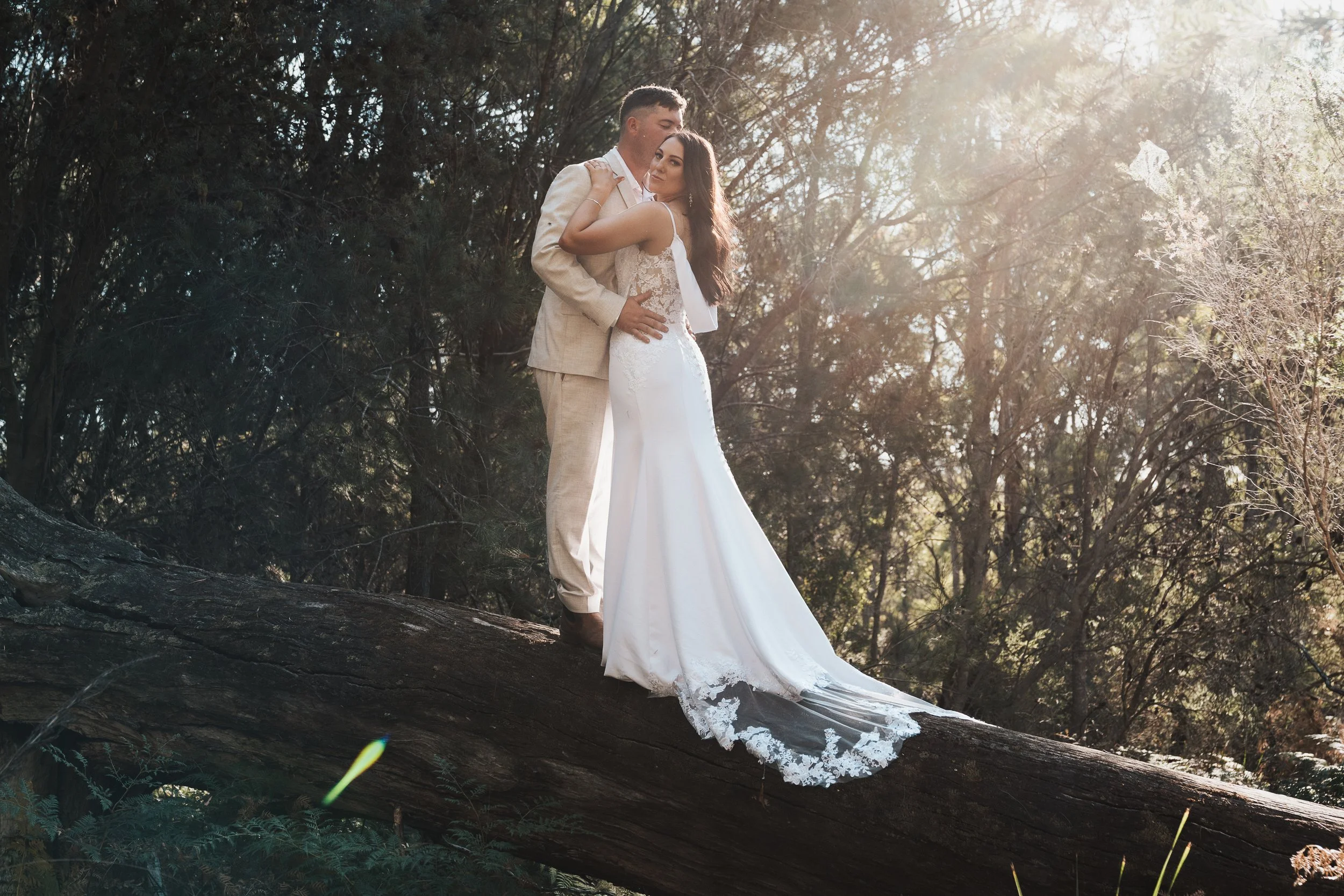 A bride and groom standing closely together outdoors on a fallen tree trunk, surrounded by trees with sunlight filtering through the branches.