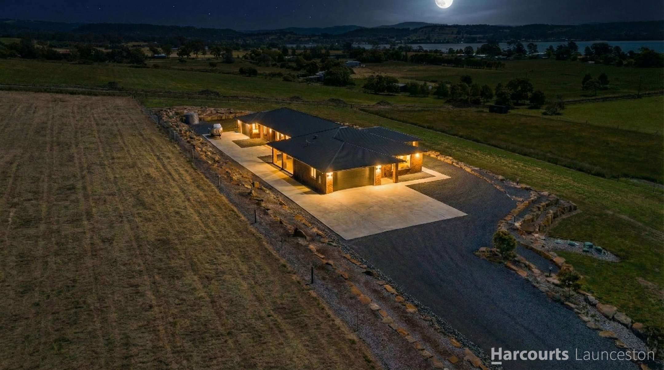 A house illuminated at night in a rural landscape with open fields, hills, trees, a water body, and a full moon in the sky.