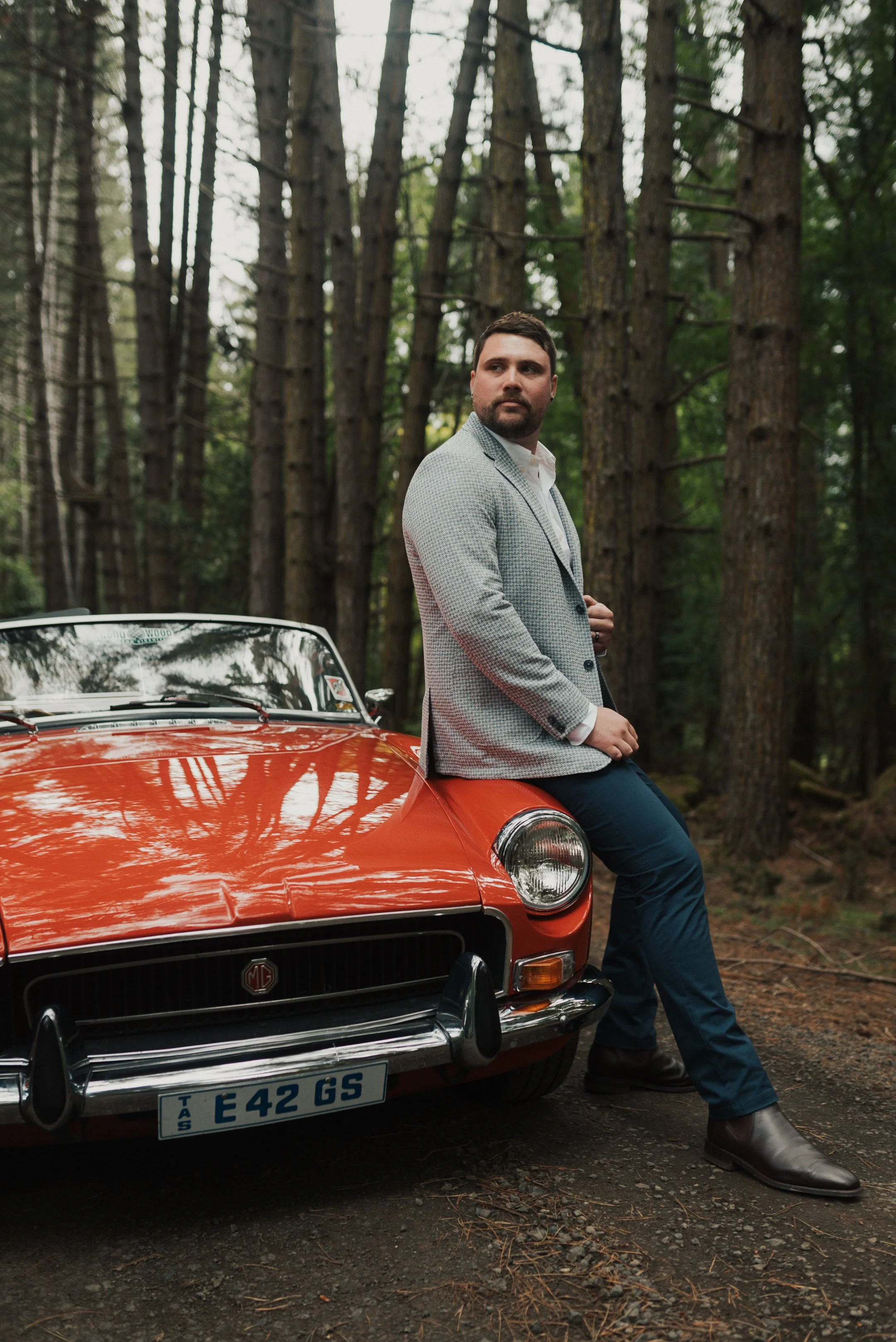 A man with a beard, dressed in a light gray blazer, white shirt, and blue pants, sitting on the front bumper of an orange vintage MG car in a wooded area.