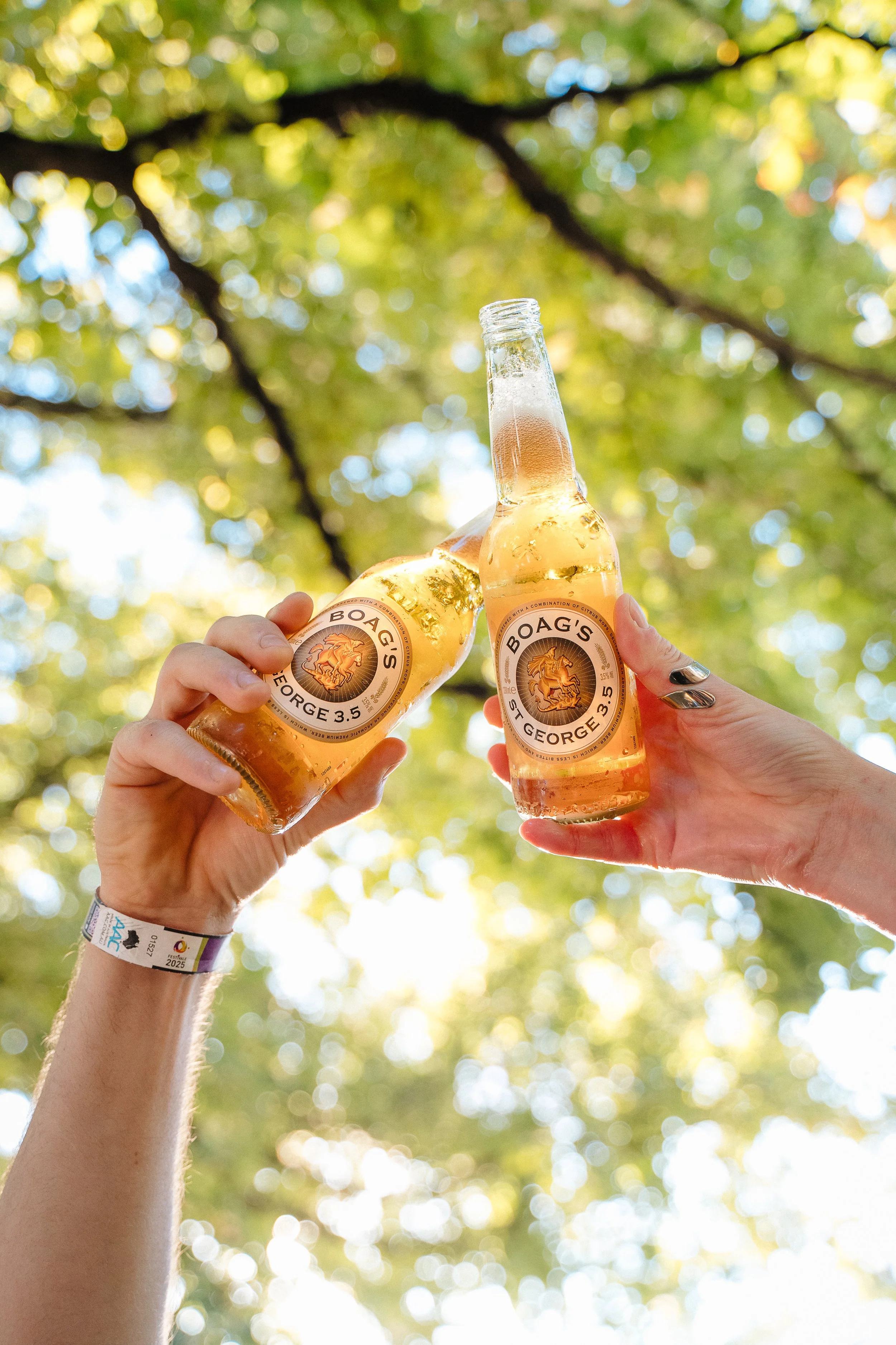 People holding up bottles of Boag's beer in an outdoor setting with leafy trees and sunlight in the background.