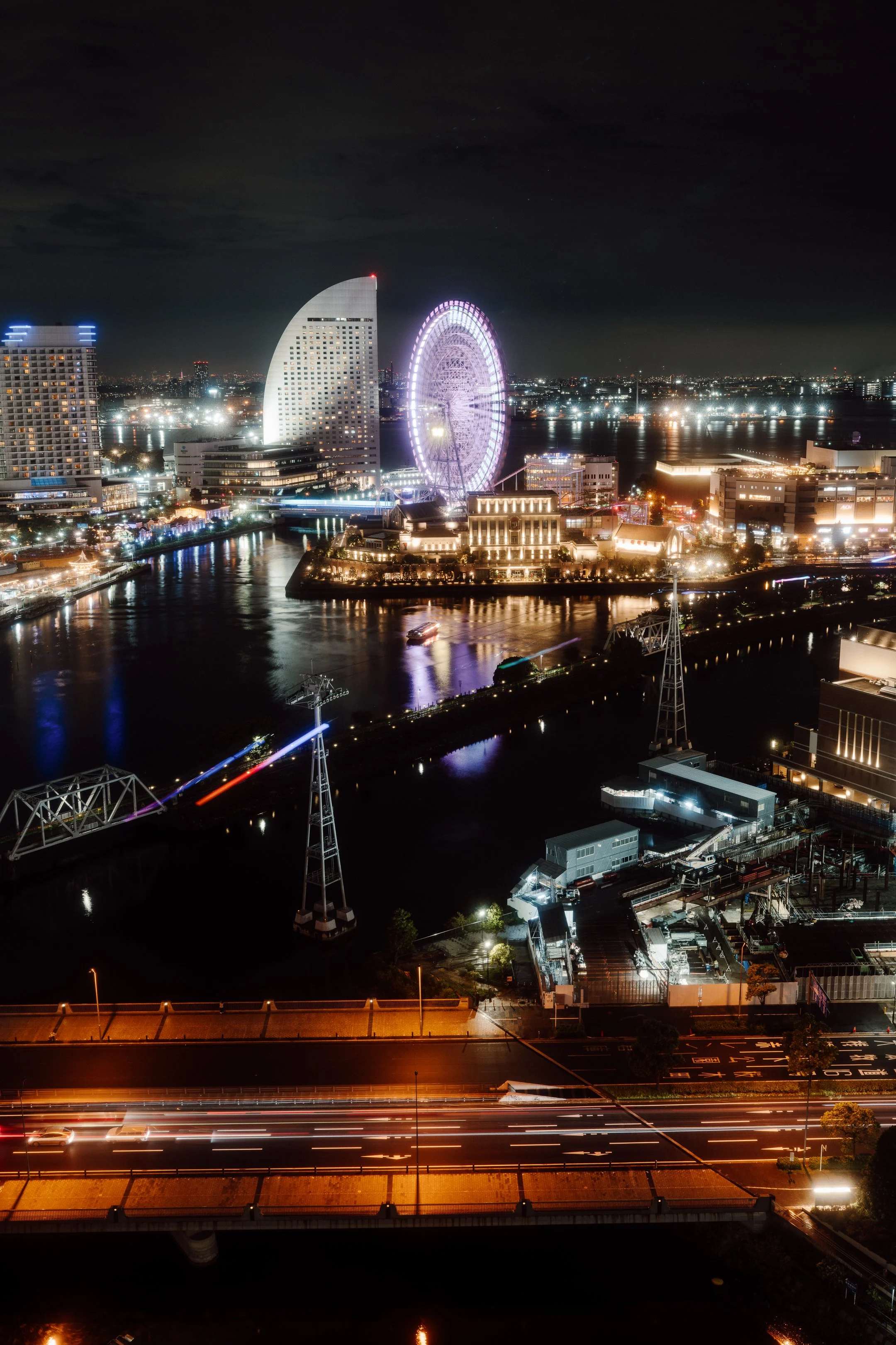 Nighttime cityscape of Yokohama, Japan, with illuminated skyscrapers, a large Ferris wheel, rivers, and light trails from moving vehicles.