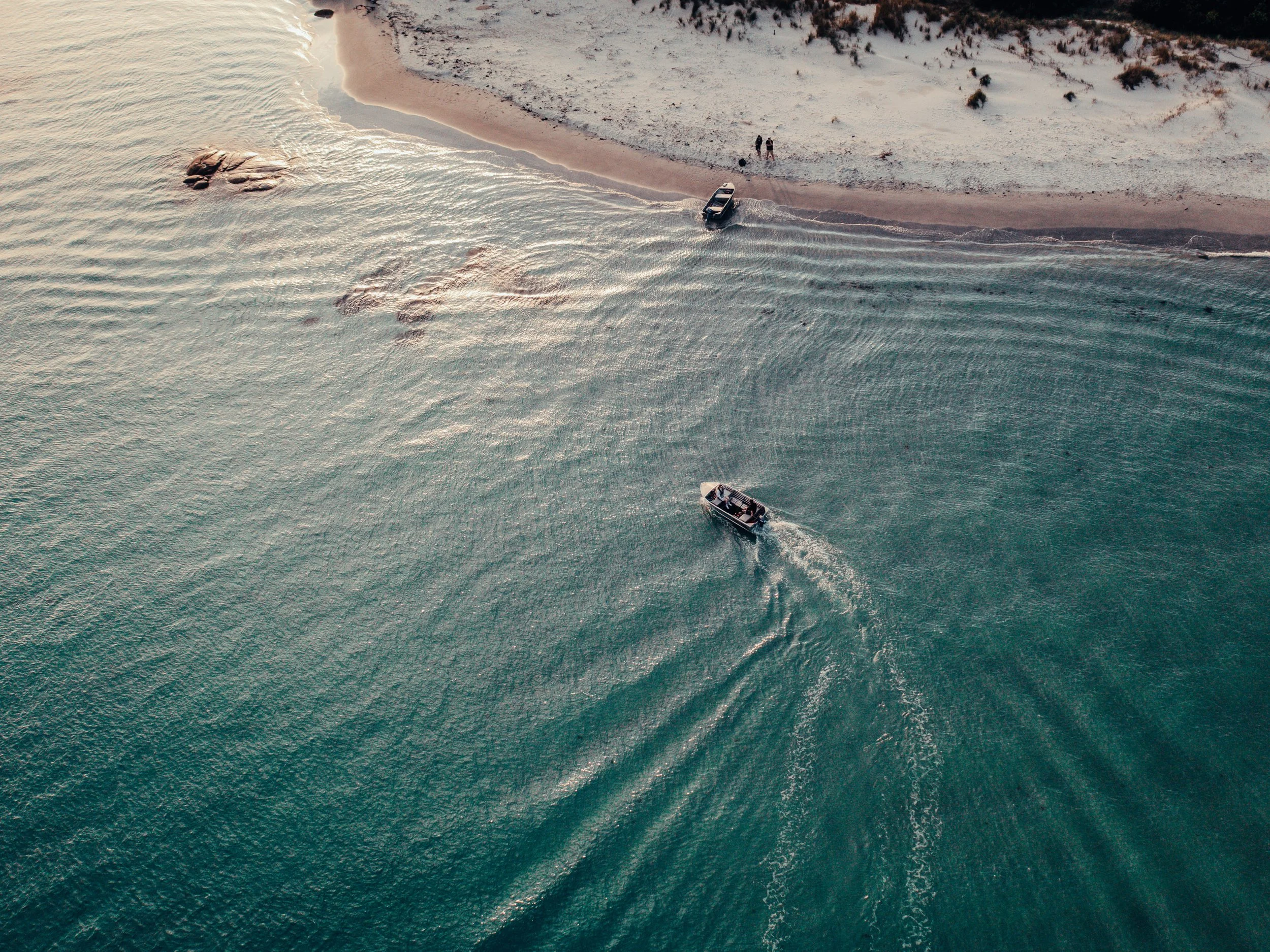 Aerial view of a boat near a sandy beach with two people standing on the shore, and rocks in the water.