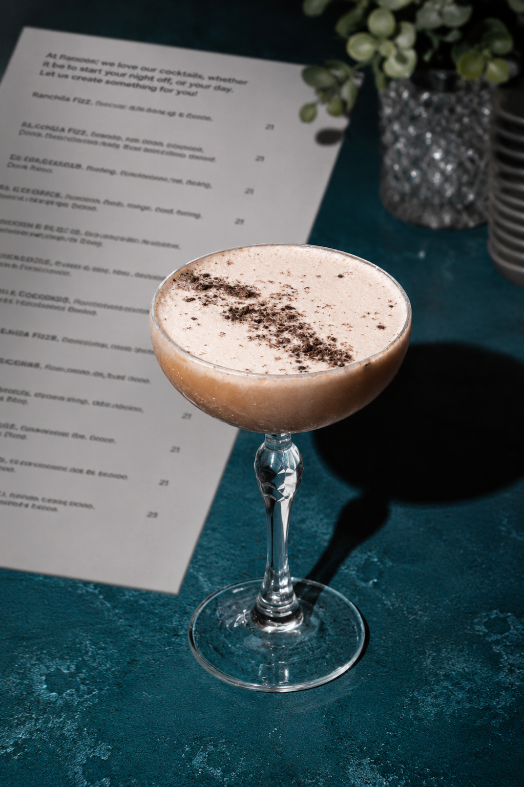 A cocktail in a coupe glass with a frothy top and chocolate shavings, placed on a textured teal surface with a menu in the background and a decorative silver vase with green leaves.
