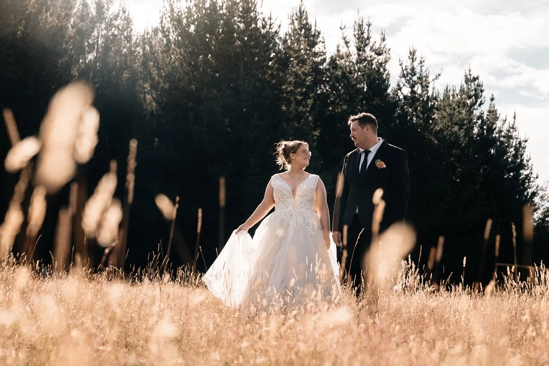 A bride and groom walking through a grassy field during sunset with a forest backdrop, smiling at each other.