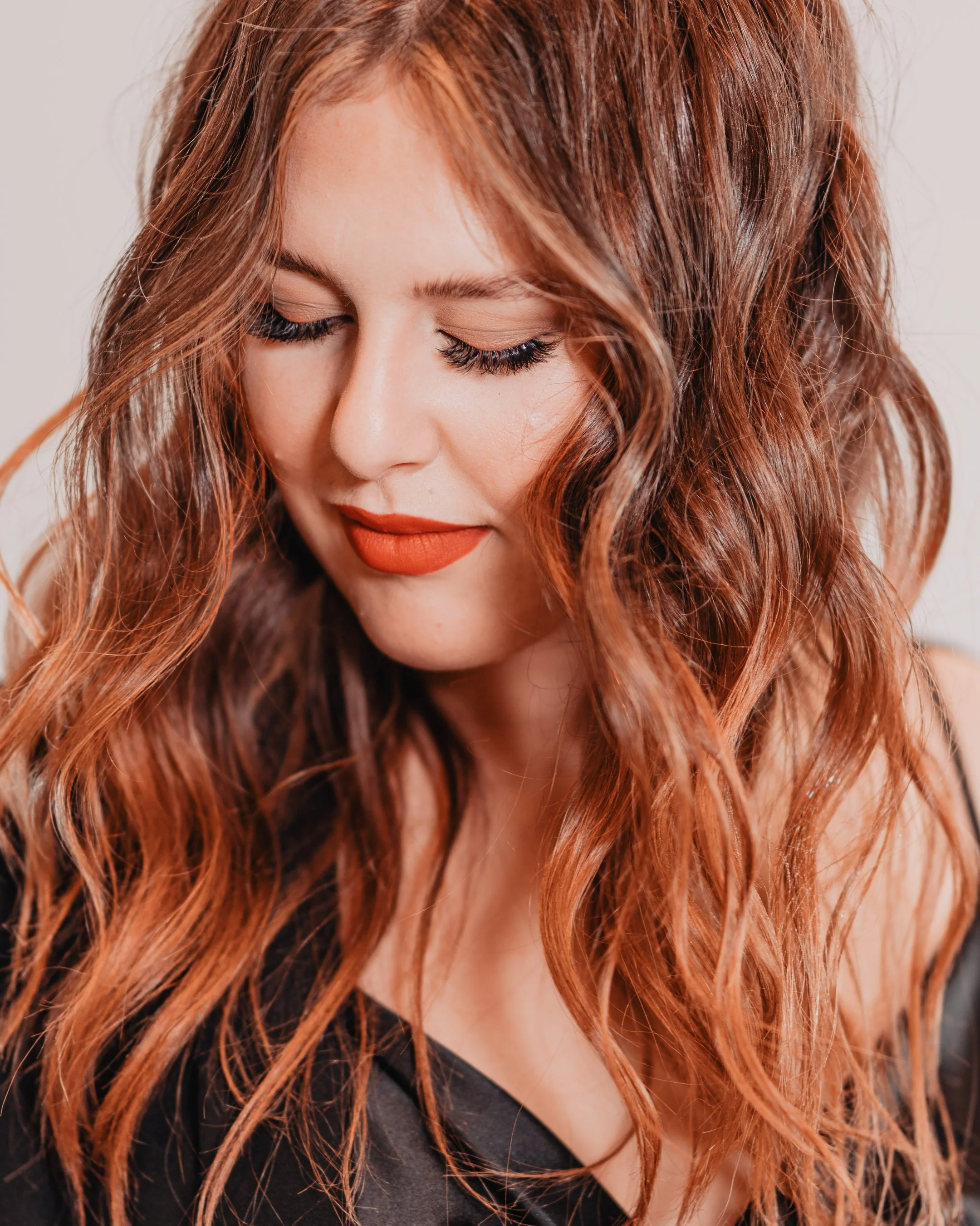 Close-up of a woman with long, wavy, red hair, wearing nude lipstick and having her eyes closed, against a plain background.