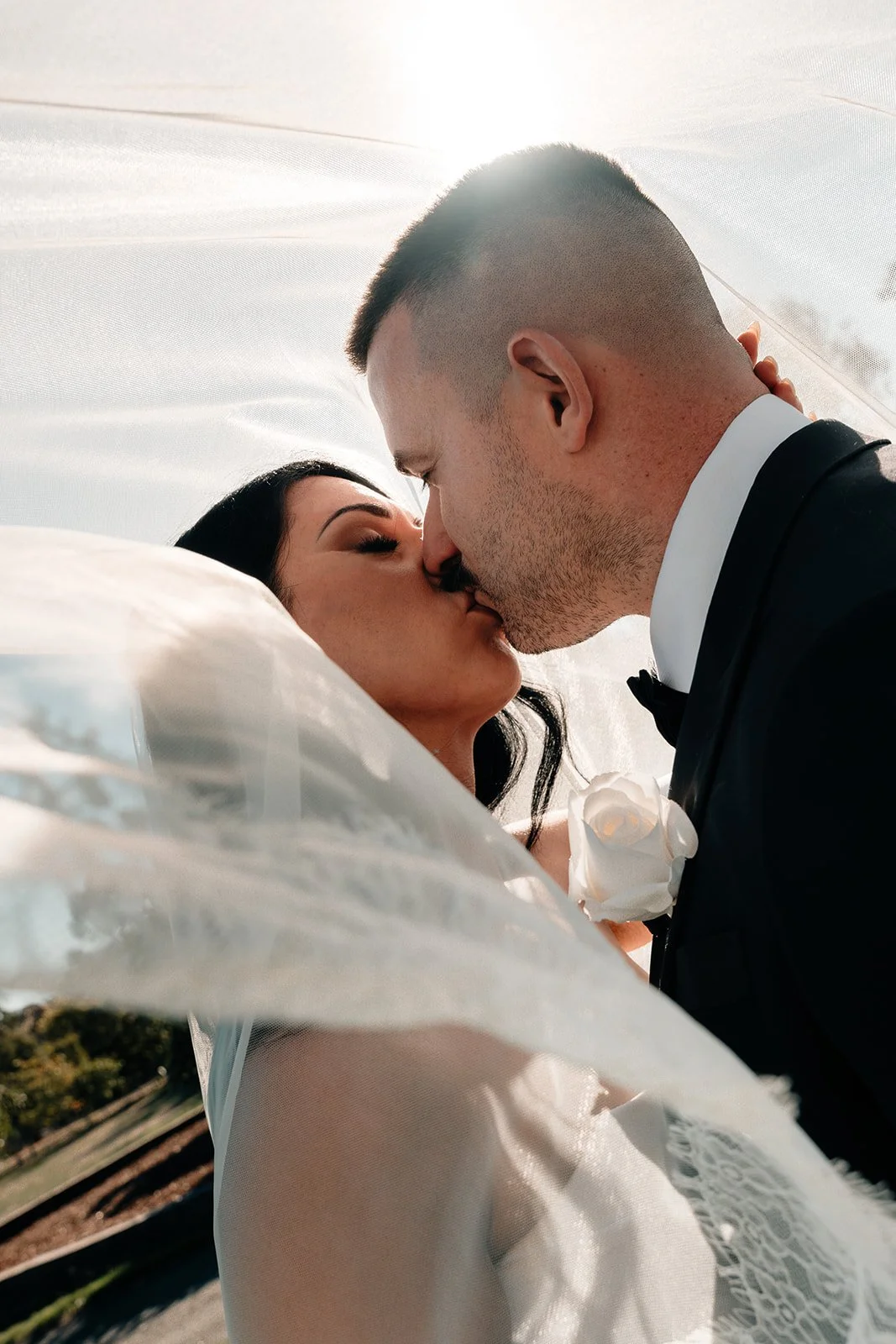 Couple kissing during wedding, bride with veil and groom in tuxedo outdoors.