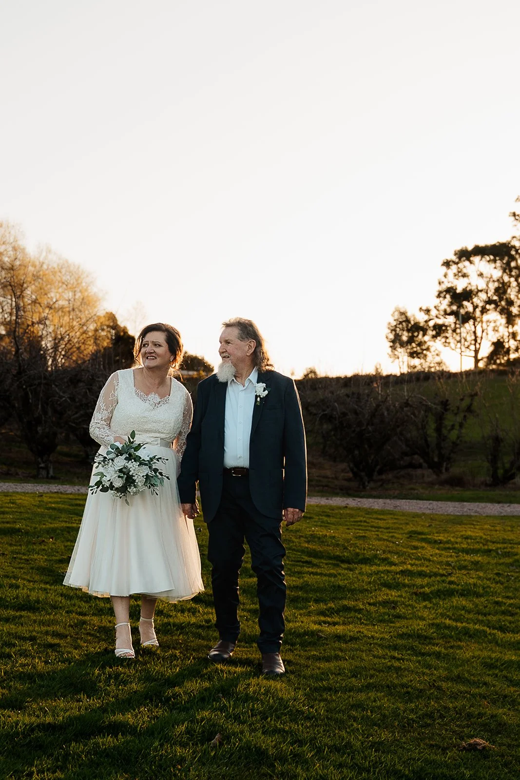 A bride in a white wedding dress holding a bouquet, walking hand-in-hand with a man in a dark suit, in an outdoor setting during sunset with trees in the background.