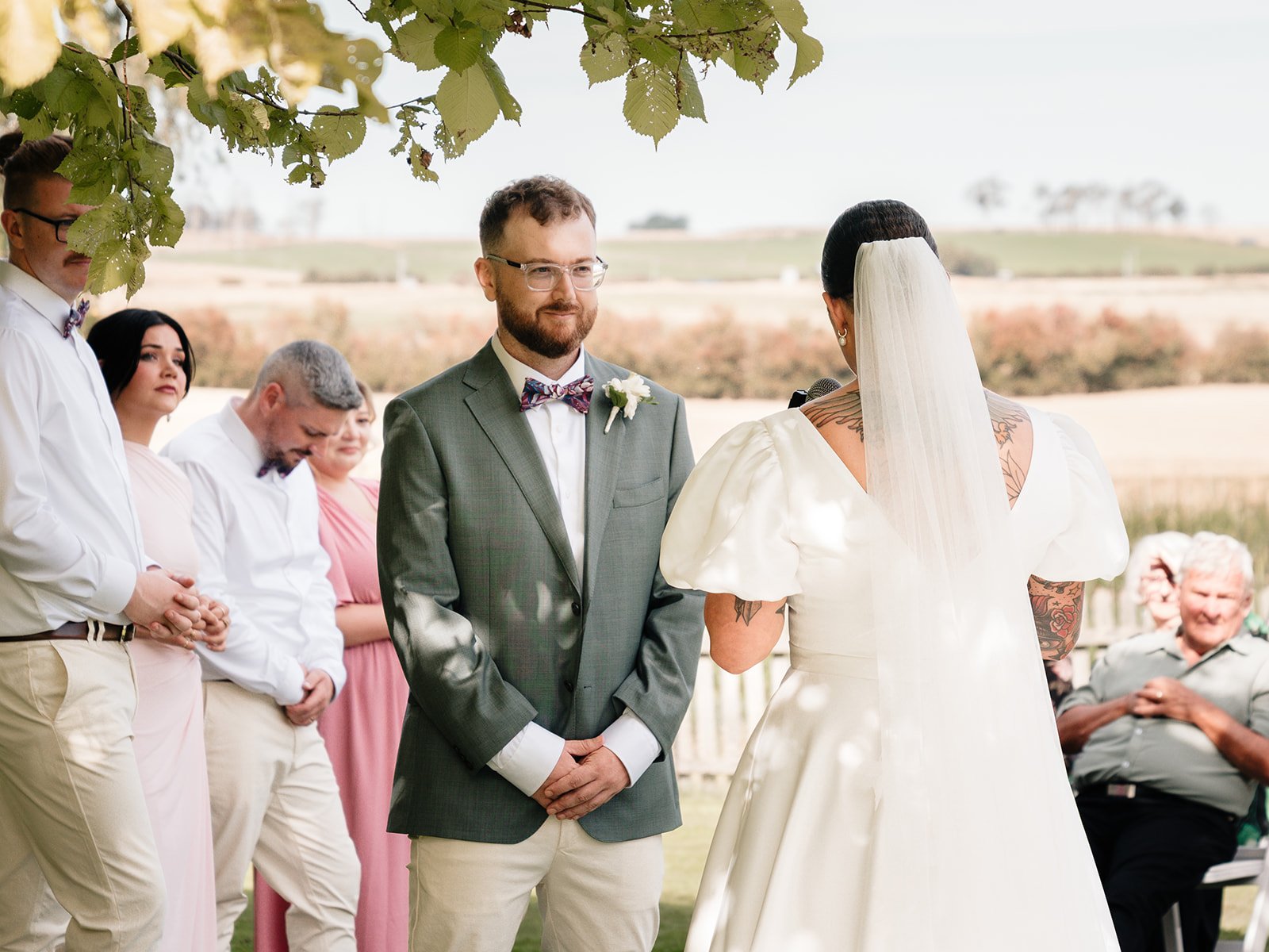 A wedding ceremony outdoors with a groom in a gray suit and glasses looking at the bride, who has tattoos and is wearing a white dress with puffed sleeves and a veil. Several guests are standing behind them, including a woman in pink and men in white