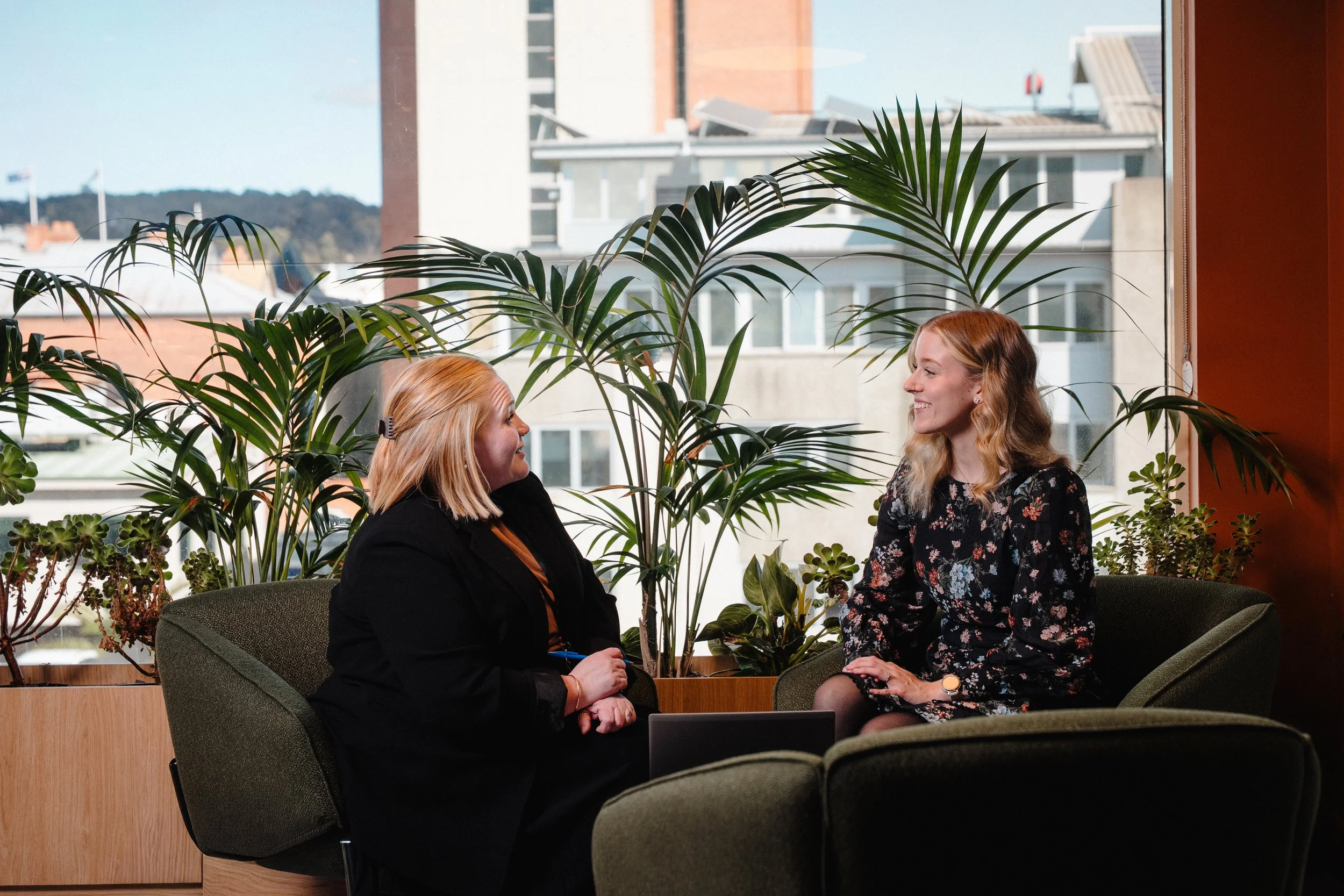 Two women sitting and conversing in an indoor space with large window, plants, and cityscape view outside.