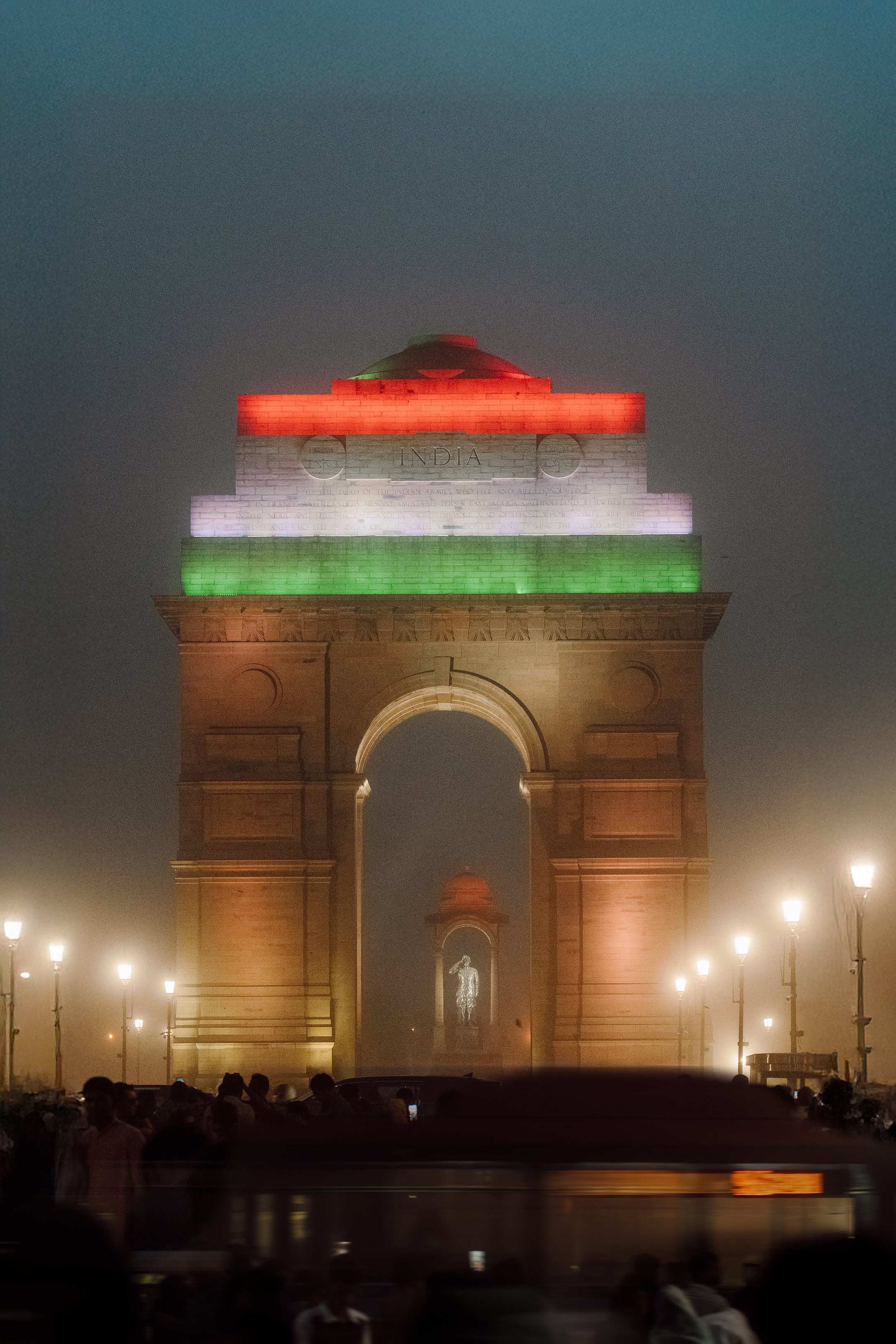 India Gate monument illuminated with the colors of the Indian flag: orange, white, and green, during the night, with people and streetlights in the surrounding area.