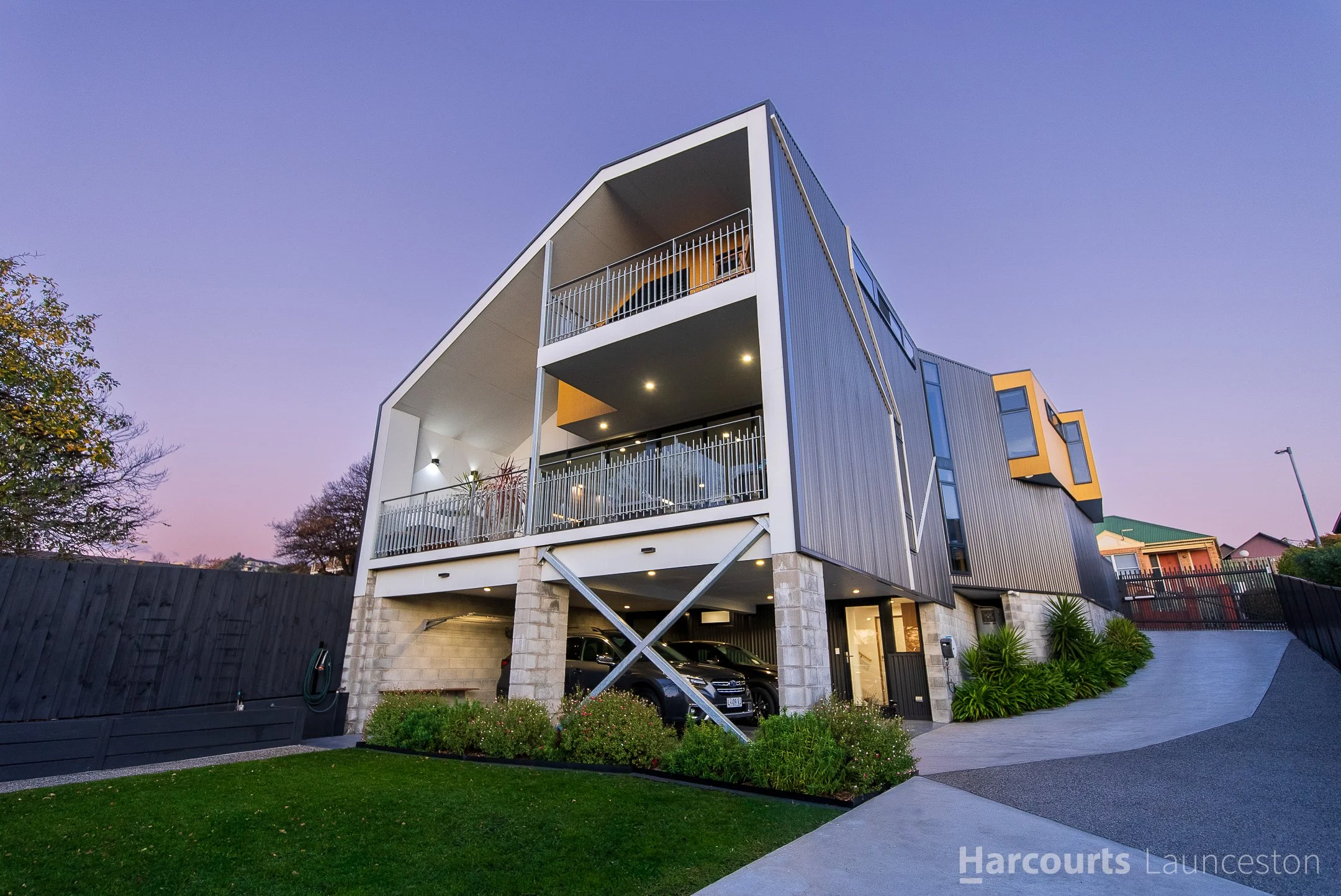Modern multi-story building with balconies, parking garage at the bottom, and landscaped yard with grass, bushes, and trees in the foreground, during twilight.