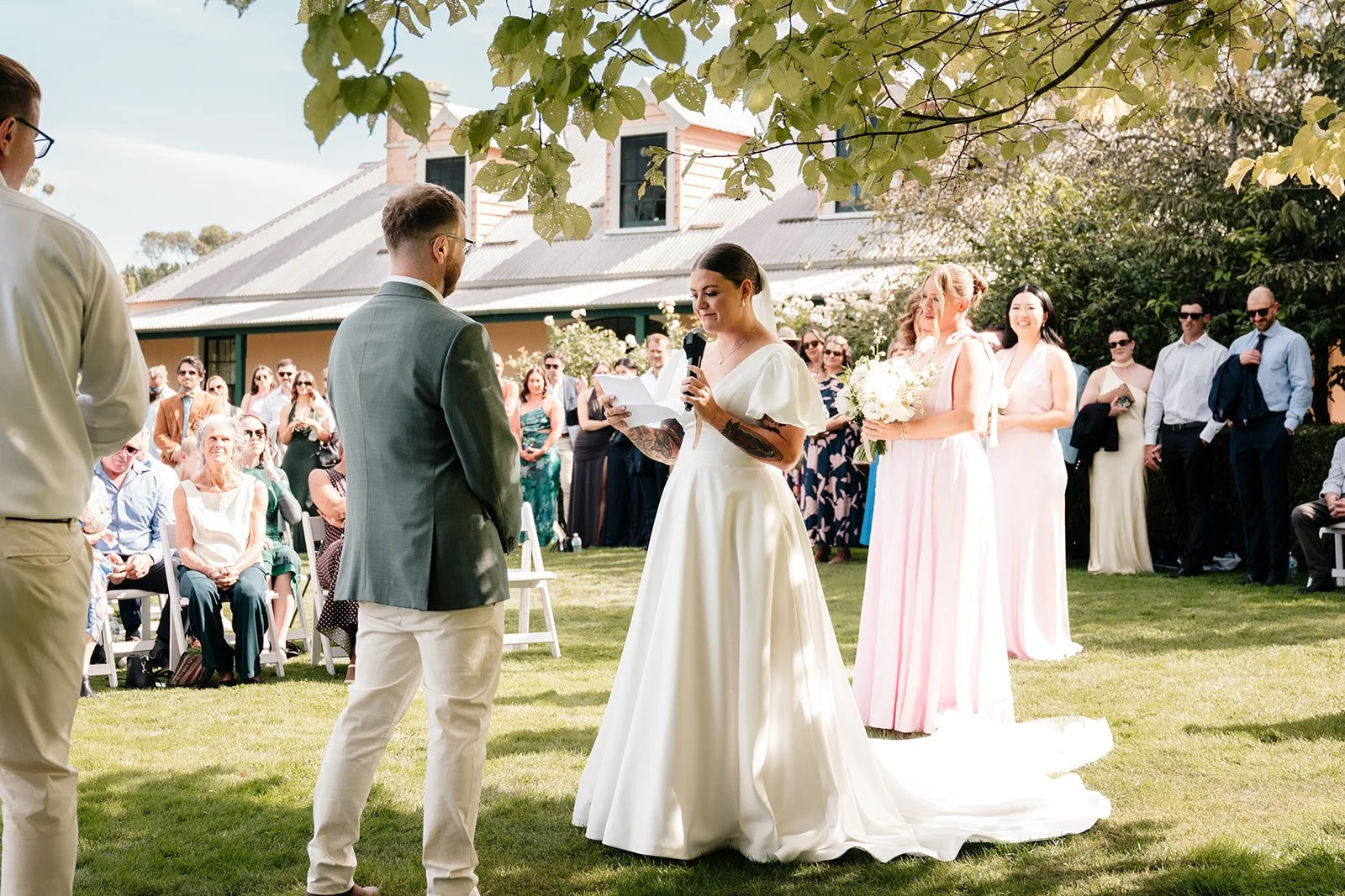 A wedding ceremony is taking place outdoors with the bride and groom exchanging vows. The bride is holding a microphone, reading her vows, and wearing a white gown. The groom is dressed in a gray jacket and cream pants. Bridesmaids in light pink dres