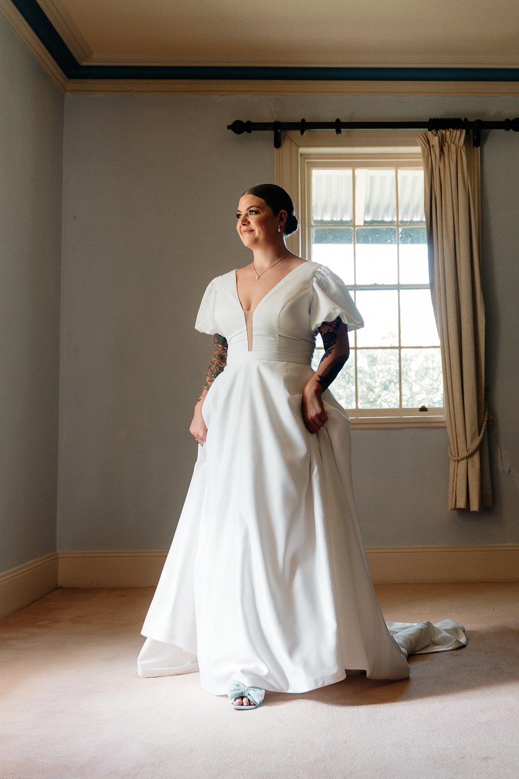 A bride in a white wedding dress standing in a room with a large window, sunlight coming through, and beige curtains, with her hands tucked into the pockets of her dress.