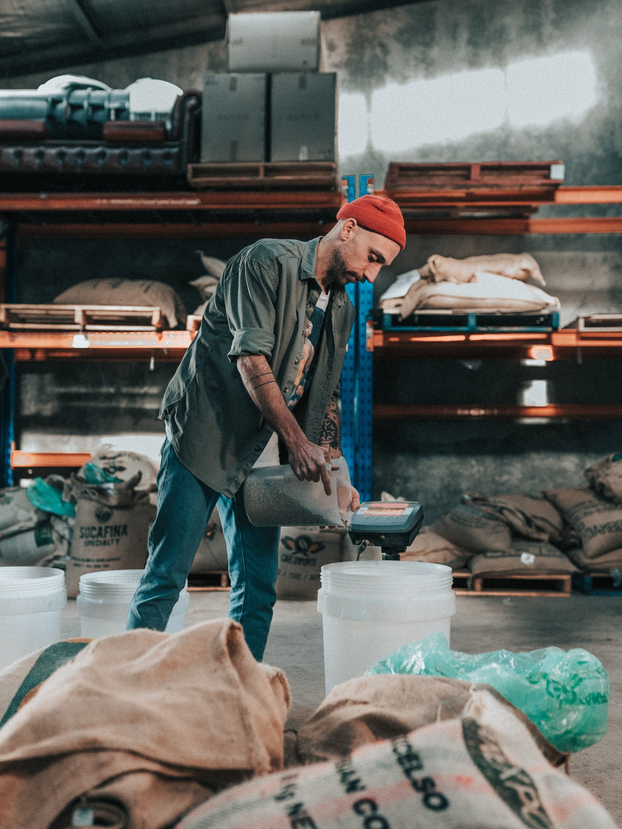 A man in a red beanie and green shirt measuring something on a device in a warehouse with shelves of bags and containers in the background.