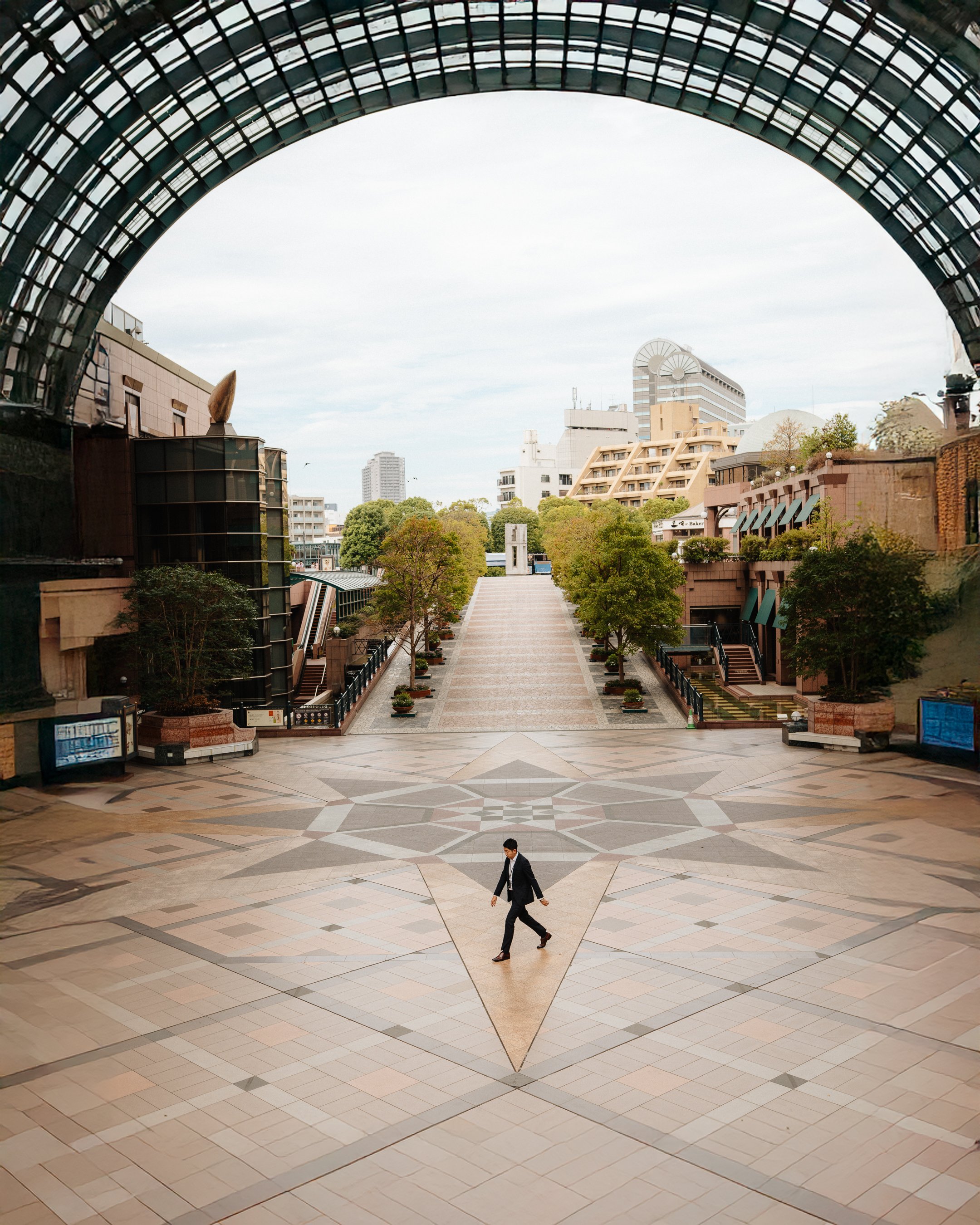 A man in a business suit walking across a large open lobby with distinctive geometric tile patterns on the floor. The background features a cityscape view through an archway, with modern buildings and trees visible outside.