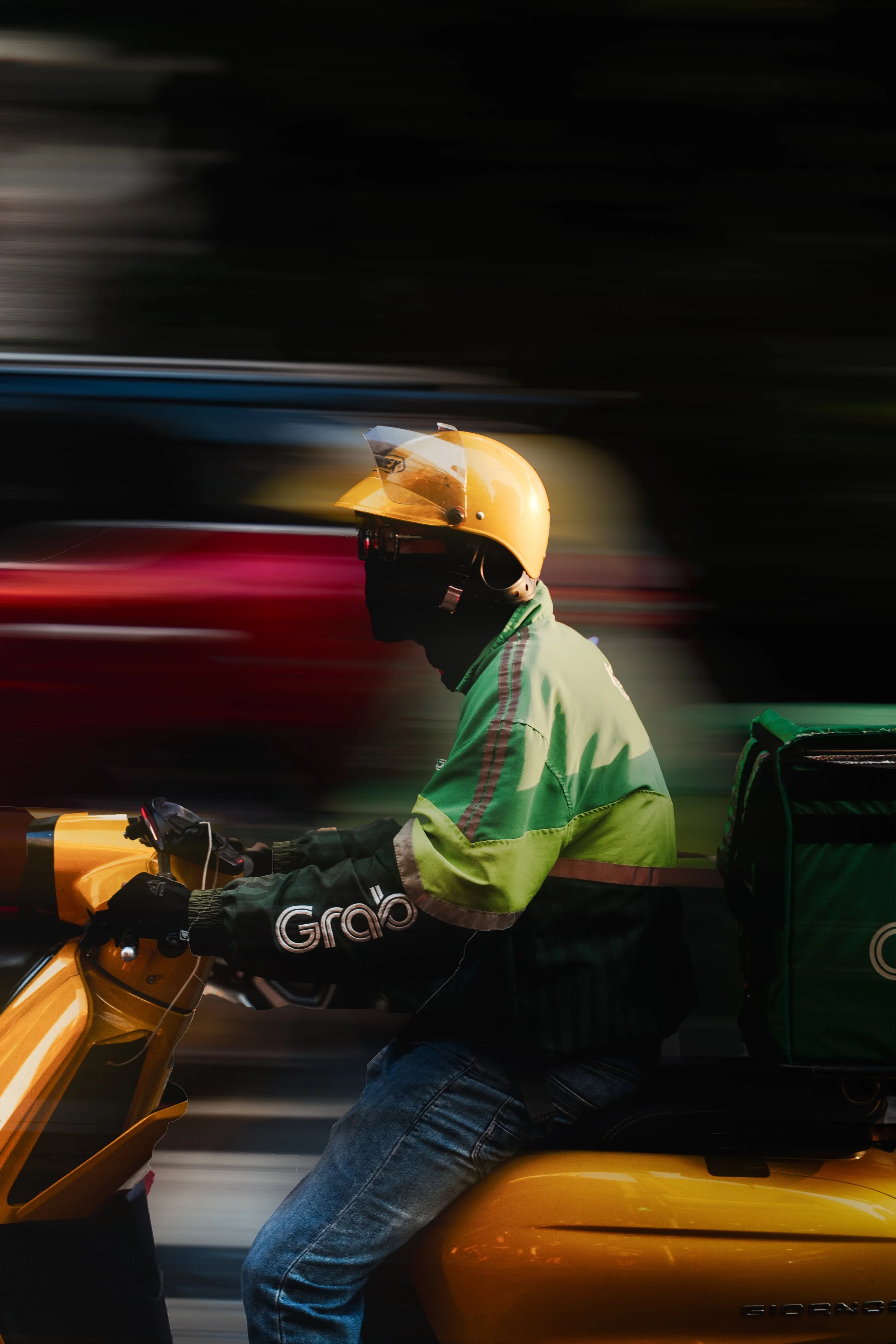 A person riding a yellow Grab scooter, wearing a yellow helmet with a visor, a green jacket, and jeans, carrying a green delivery box.