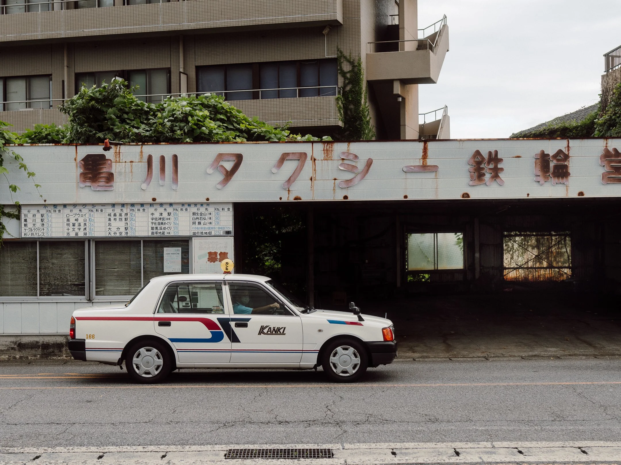 A white car with blue and red stripes driving on the street in front of a building with Japanese characters on a sign and a bus timetable. There are plants and a building with multiple balconies in the background.