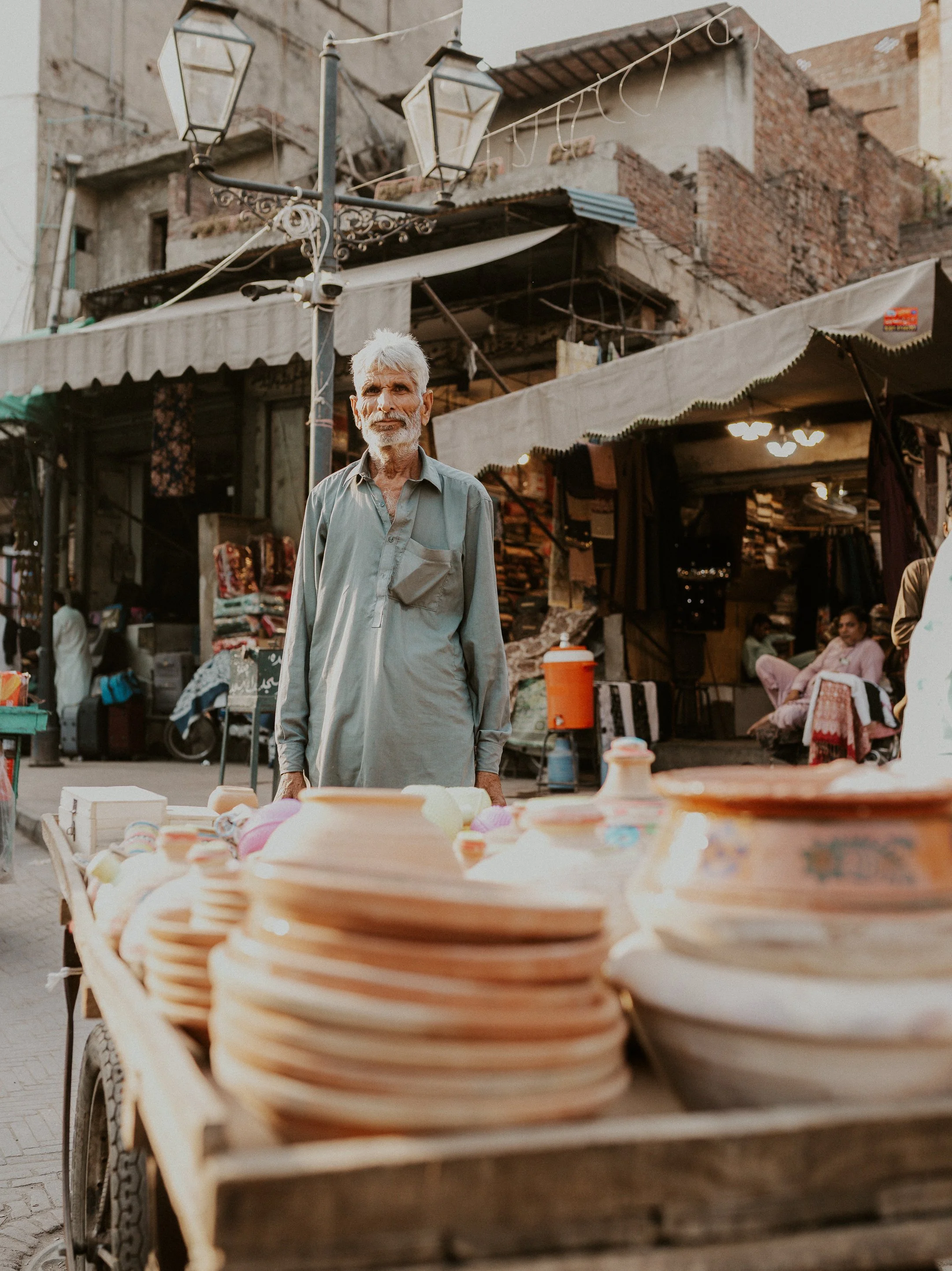 A man with gray hair and a beard stands behind a cart of ceramic plates in an outdoor market. In the background, there are buildings with exposed brick and fabric canopies, some people sitting and walking.