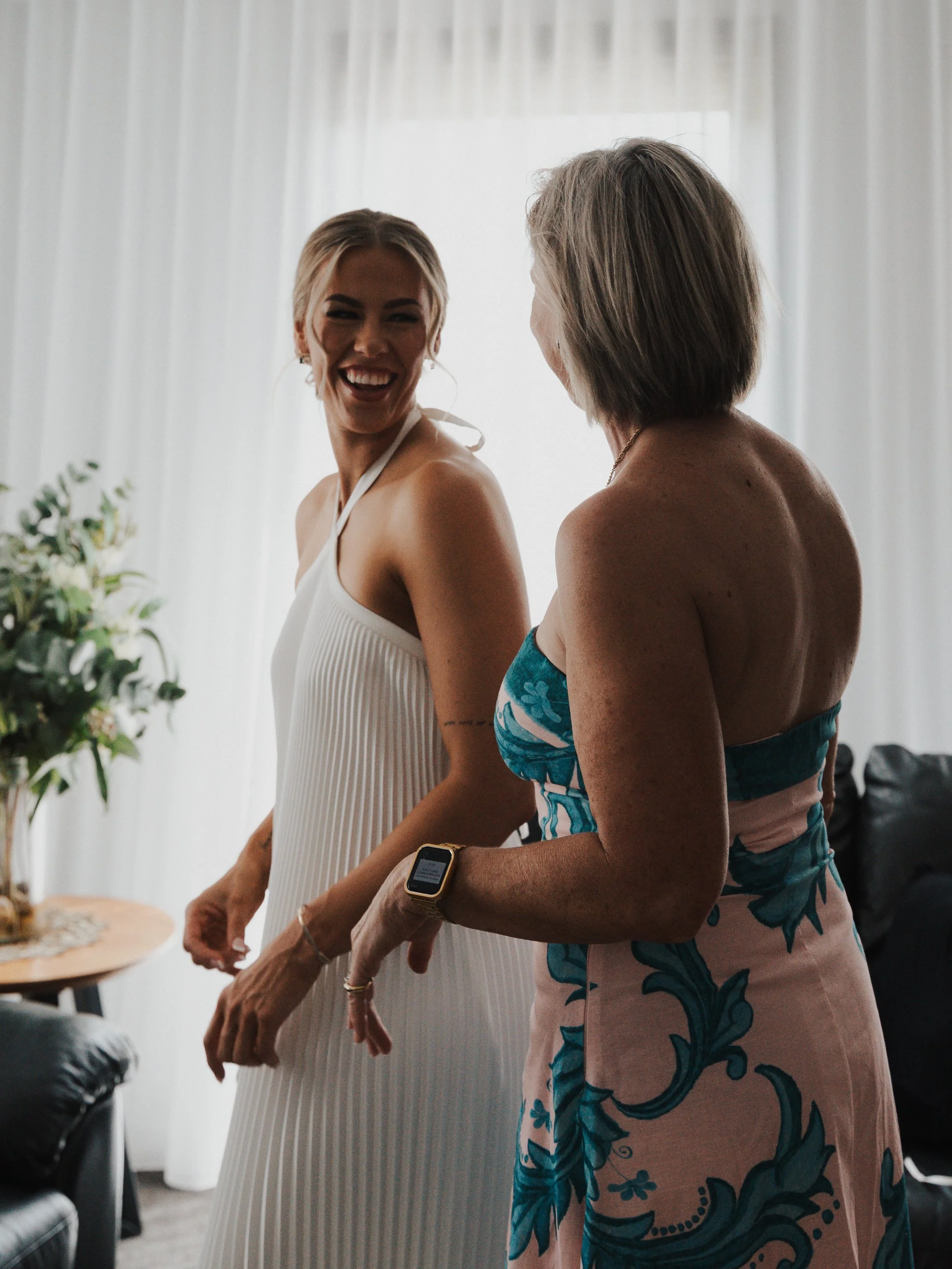 Two women smiling and talking in a living room, one with blonde hair wearing a white dress and the other with short hair wearing a strapless dress with a blue floral pattern.