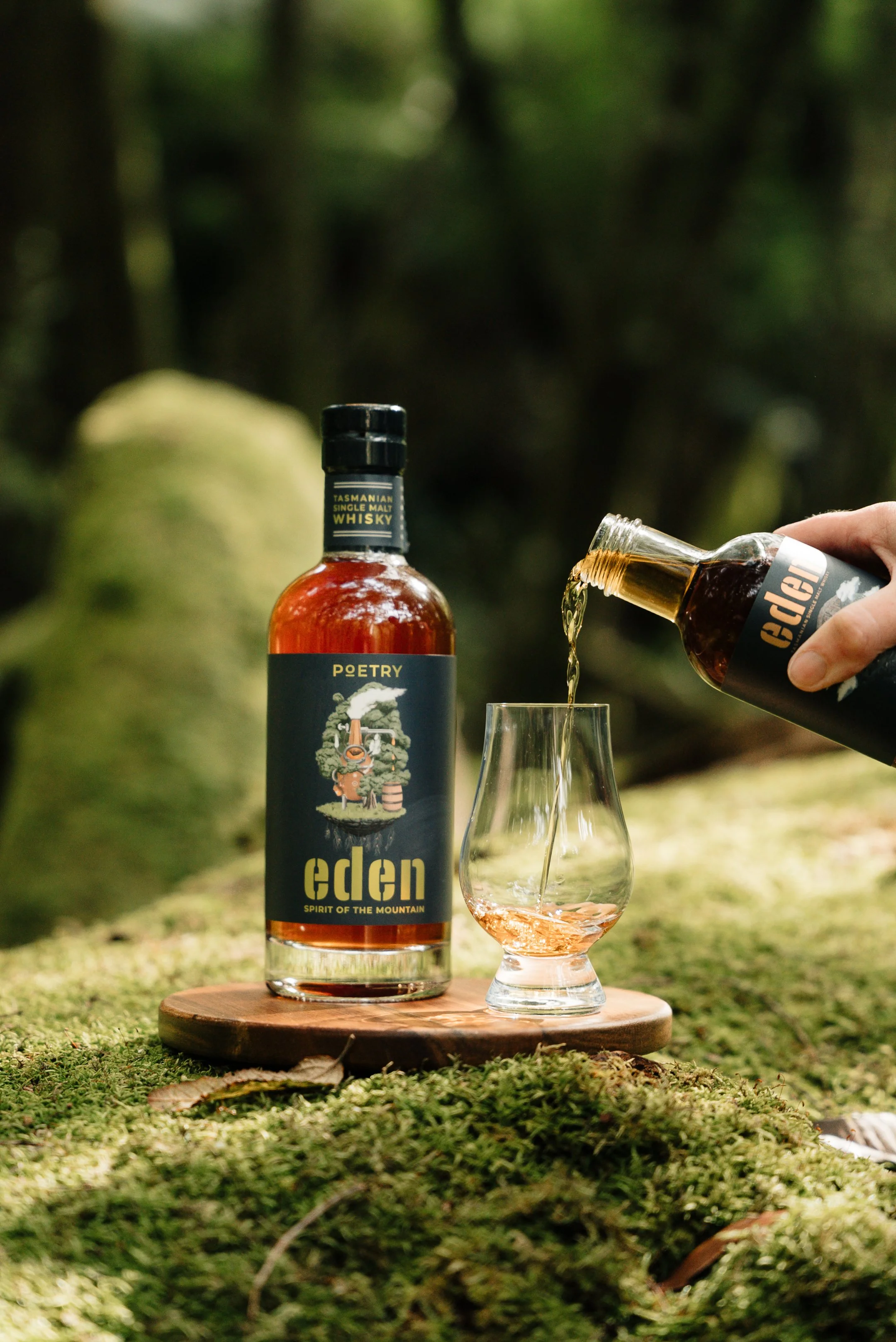 A bottle of Eden single malt whisky on a wooden board with a glass of whisky being poured into it by a person outdoors in a forest setting.