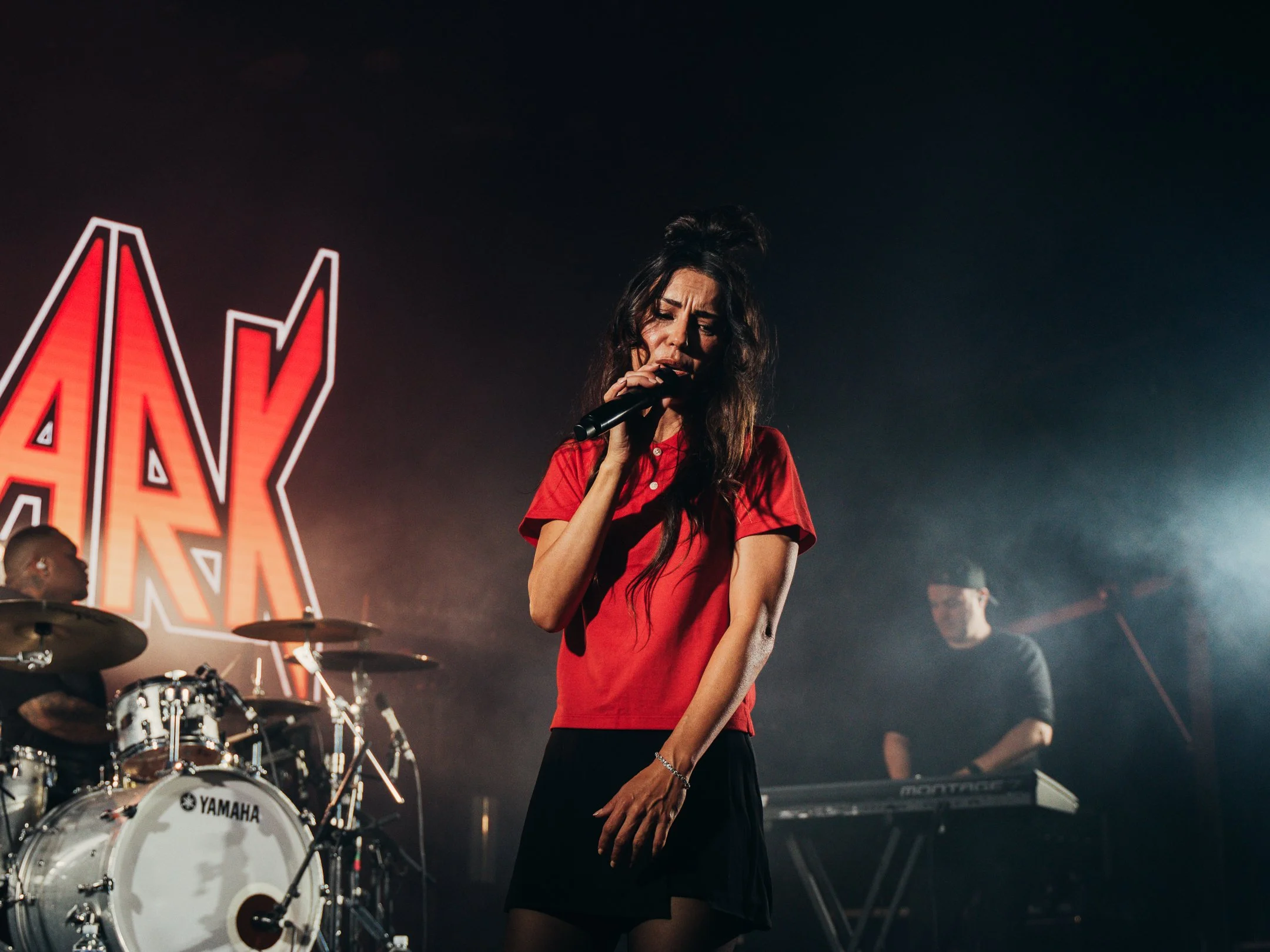 A woman singer in a red shirt performing on stage with a microphone. There are musicians and a drum set behind her, with a dark background and illuminated stage lights.