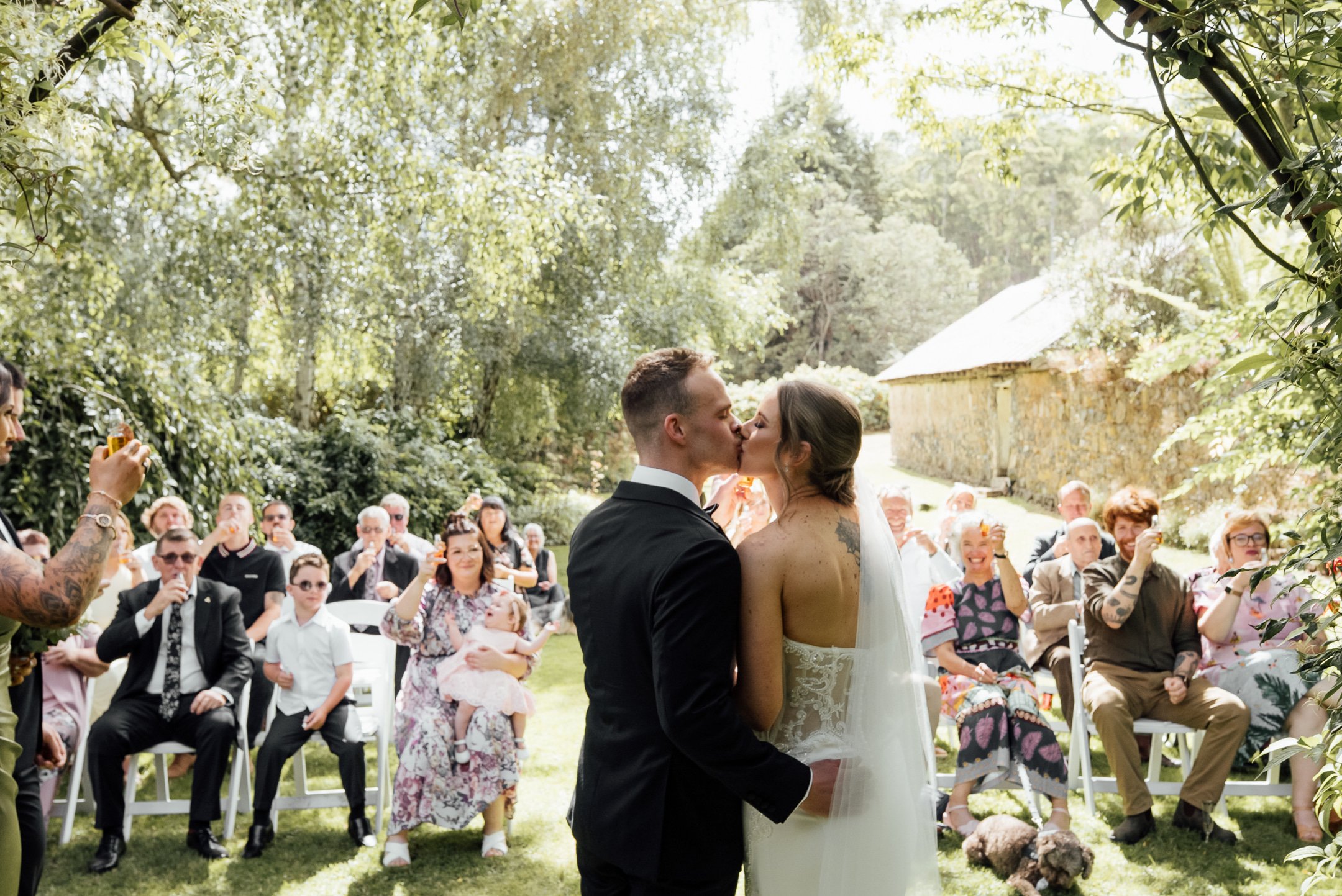 A bride and groom kiss during their outdoor wedding ceremony surrounded by guests in a lush, green setting.