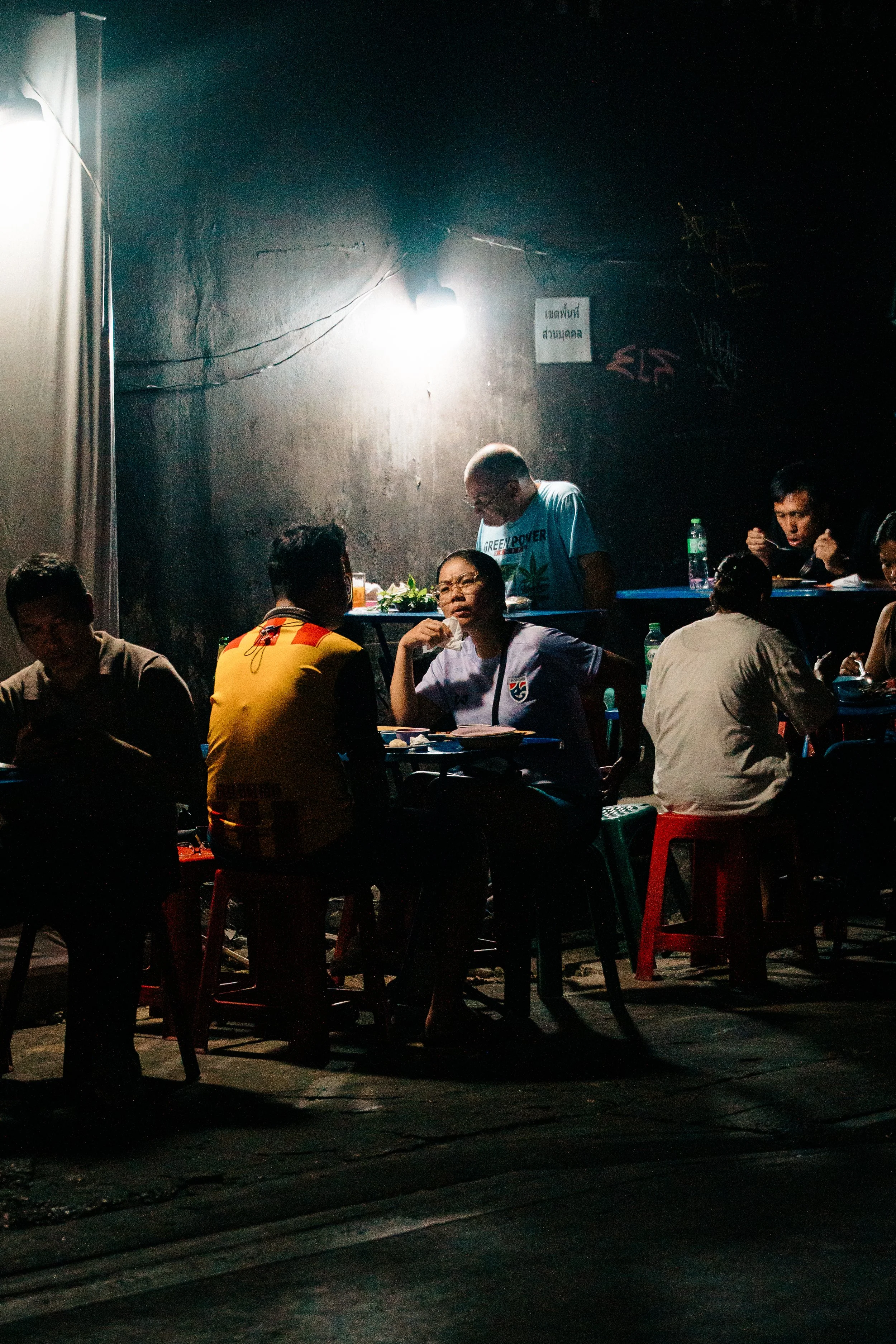 People sitting around tables outside at night, illuminated by a bright light, with a dark wall and graffiti in the background.