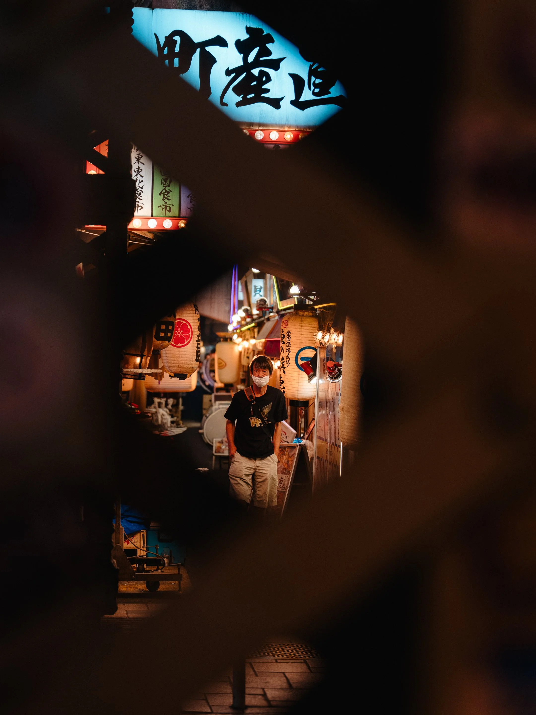 A person wearing a mask and black t-shirt standing at a night market stall with paper lanterns and colorful signs, seen through a hole in a fence or barrier.