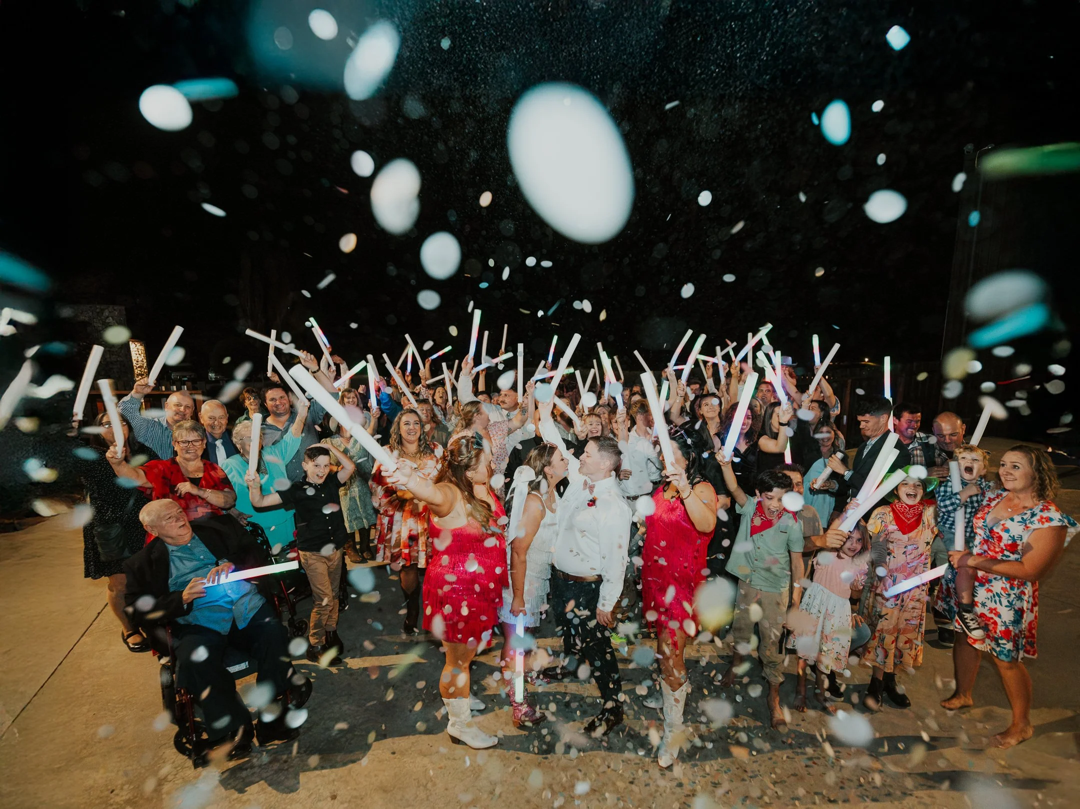 A large crowd of people celebrating at night, holding glow sticks and surrounded by falling confetti.