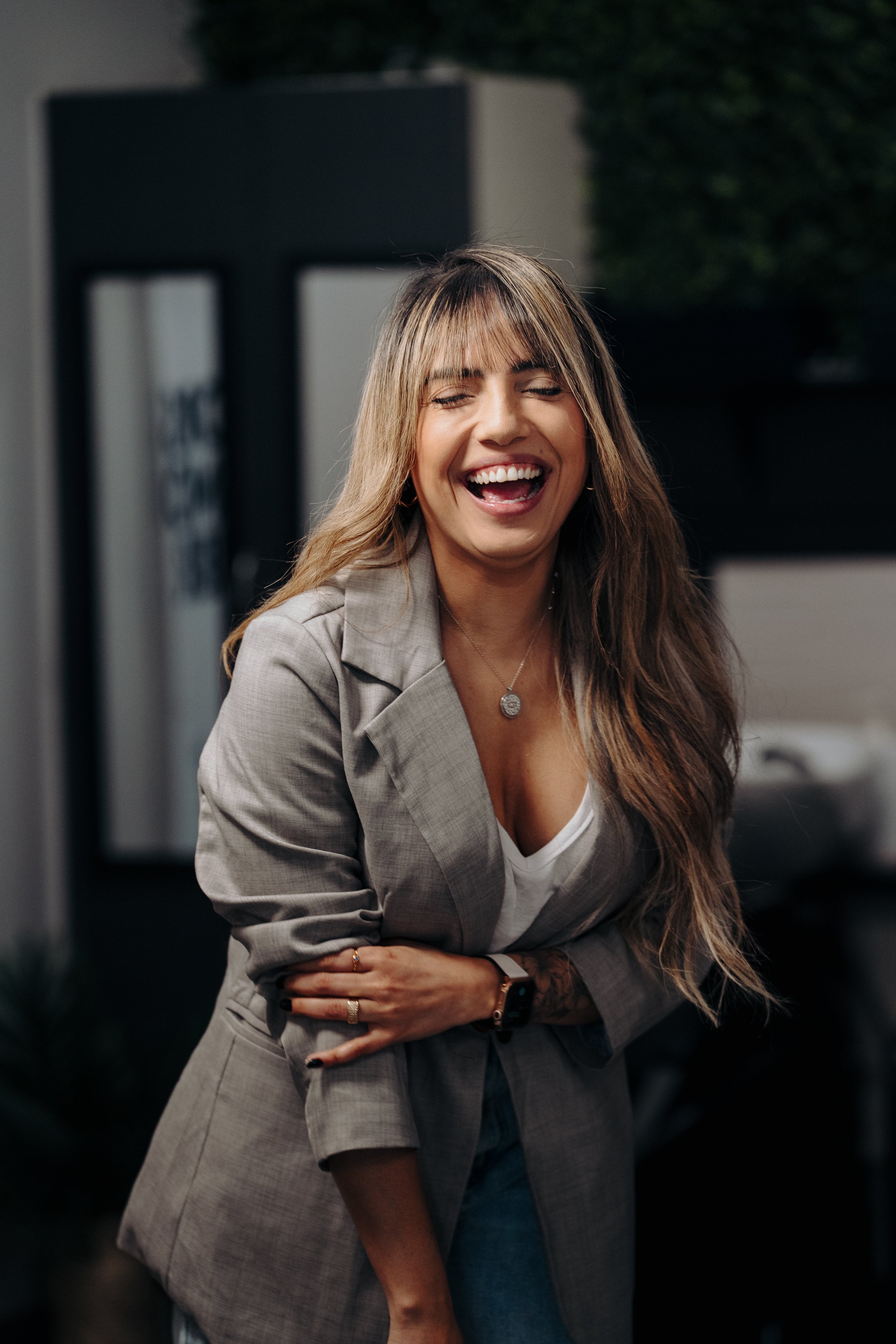 A woman with long brown hair, wearing a gray blazer and white top, laughing with eyes closed, standing indoors with a blurred dark background.