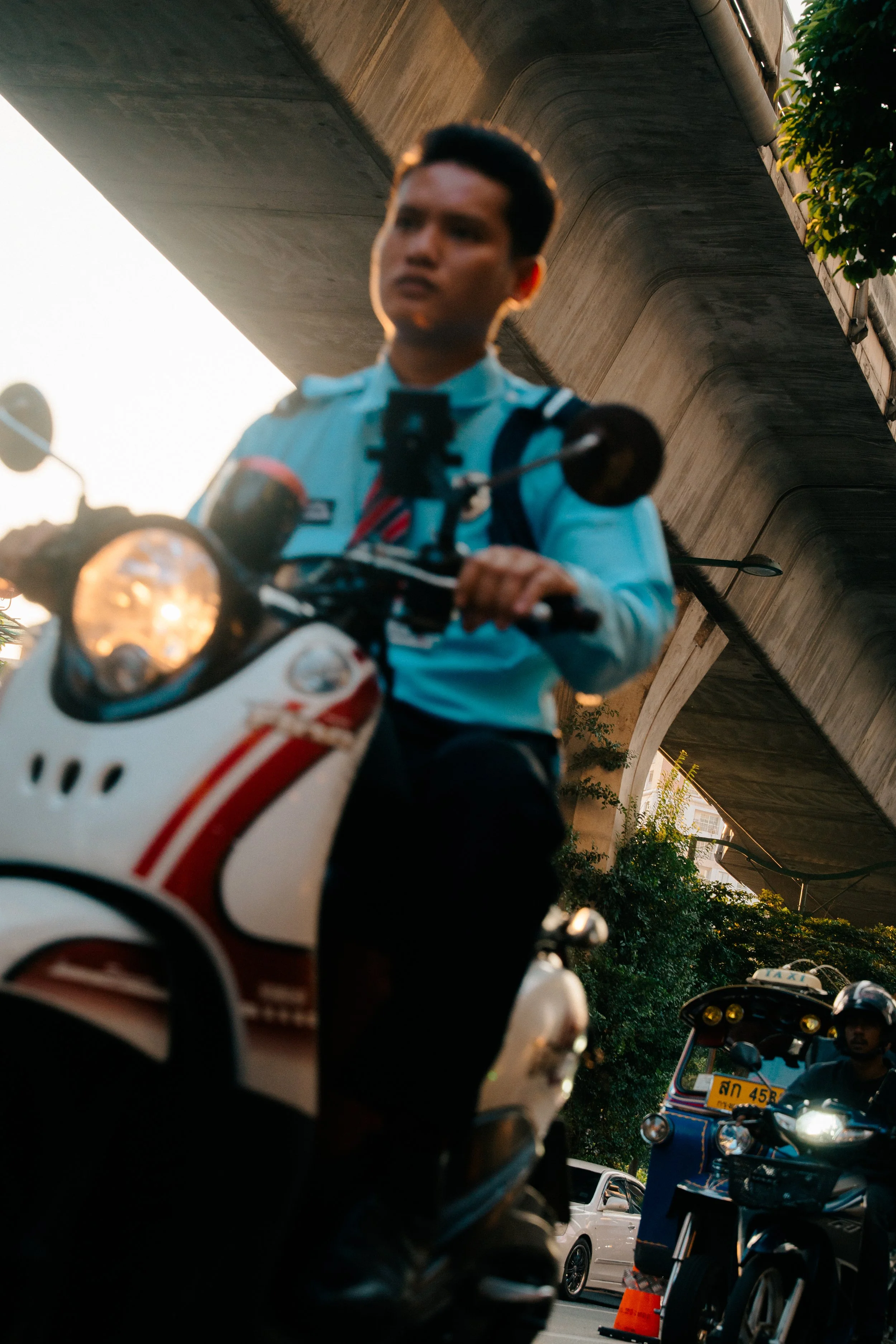 A man riding a white and red motorbike, with other vehicles around him, under a concrete bridge, during sunset.