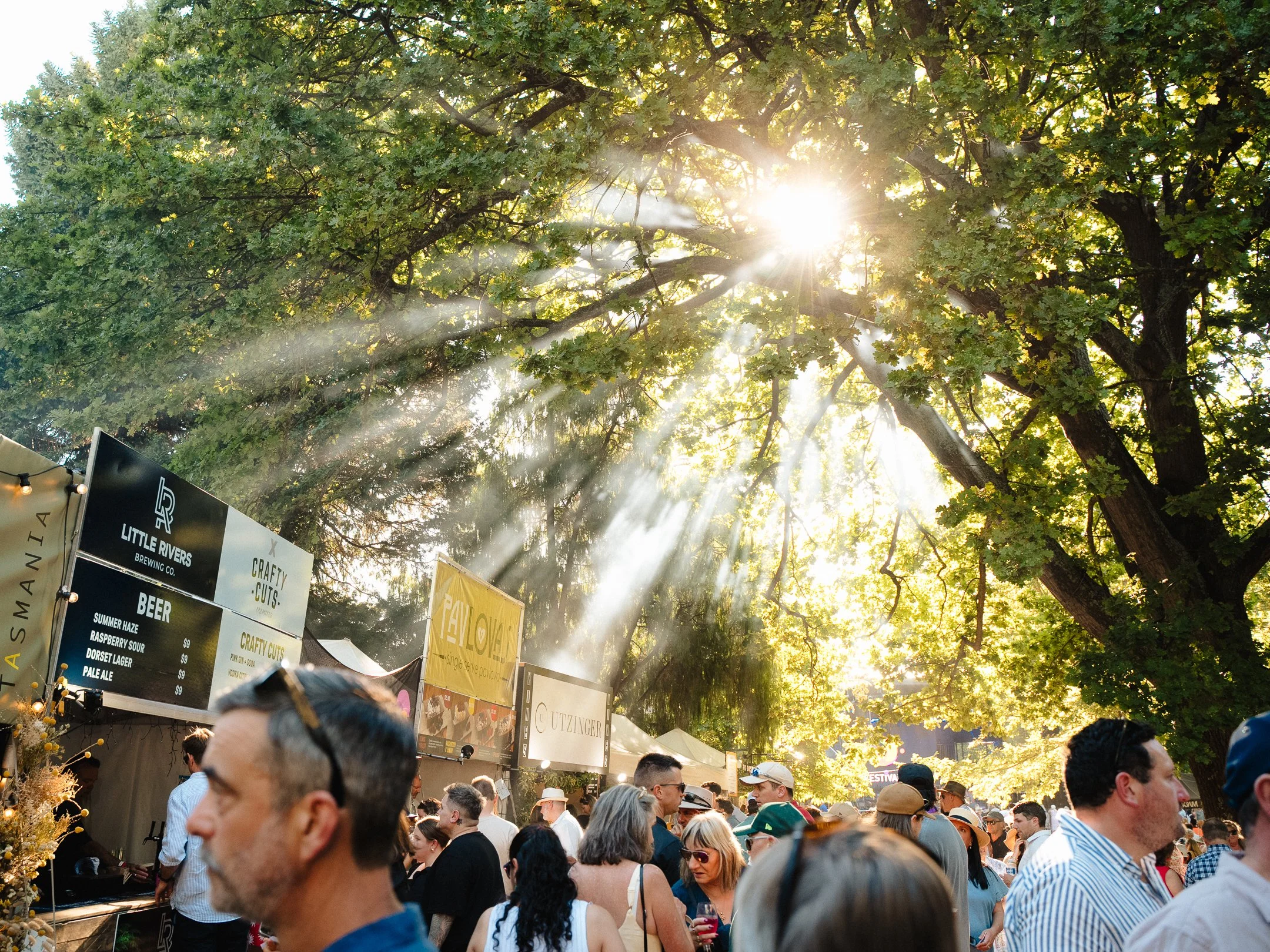 A crowded outdoor festival or market on a sunny day with sunlight streaming through green leafy trees. People are gathered around food and drink stands, with some holding drinks and chatting. Several banners and signs are visible, advertising beer and craft items.