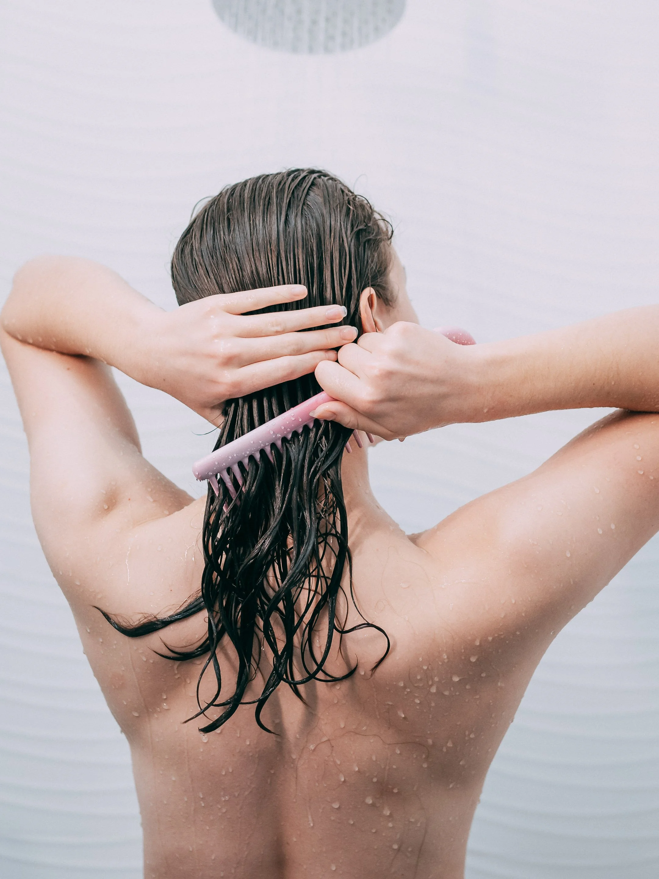 A woman with wet hair, holding a pink comb, is taking a shower under a round shower head.