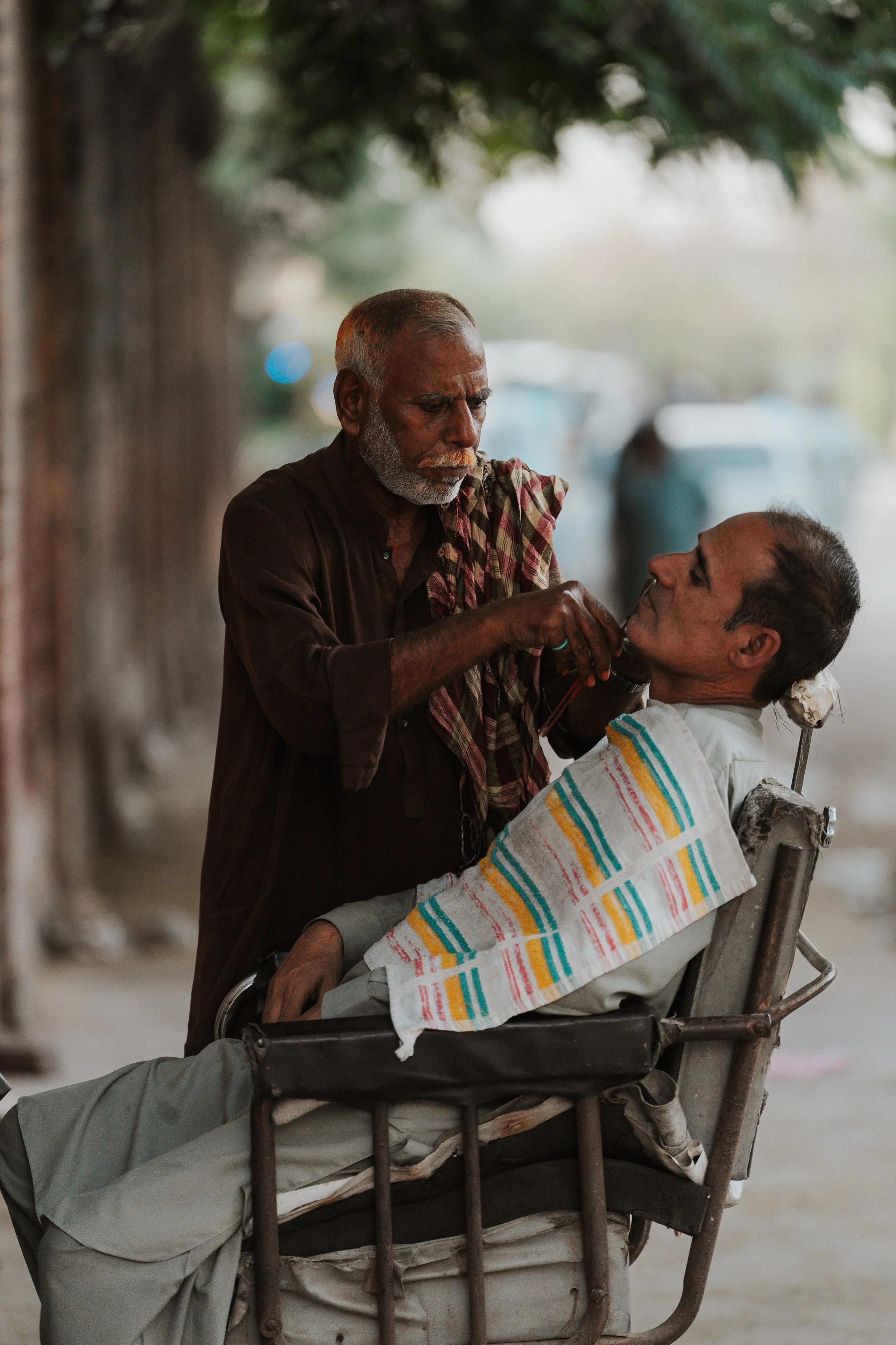 An elderly man with a gray beard is attending to a young man in a wheelchair, who is covered with a striped towel. They are outdoors under some trees, with vehicles and a blurred background visible.