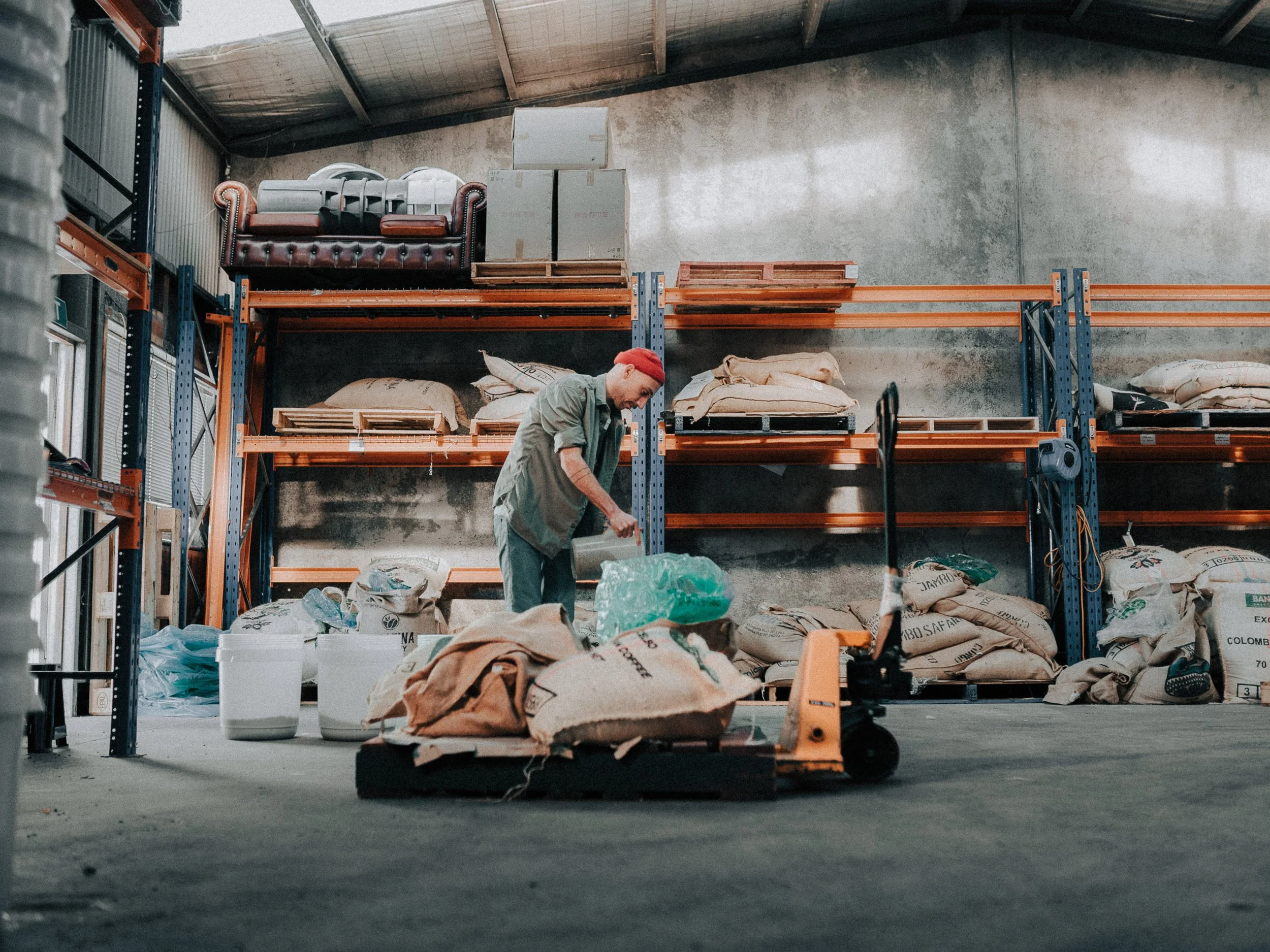 Man in a warehouse or storage facility with shelves filled with large bags, boxes, and furniture pieces, including a sofa on the upper shelf.