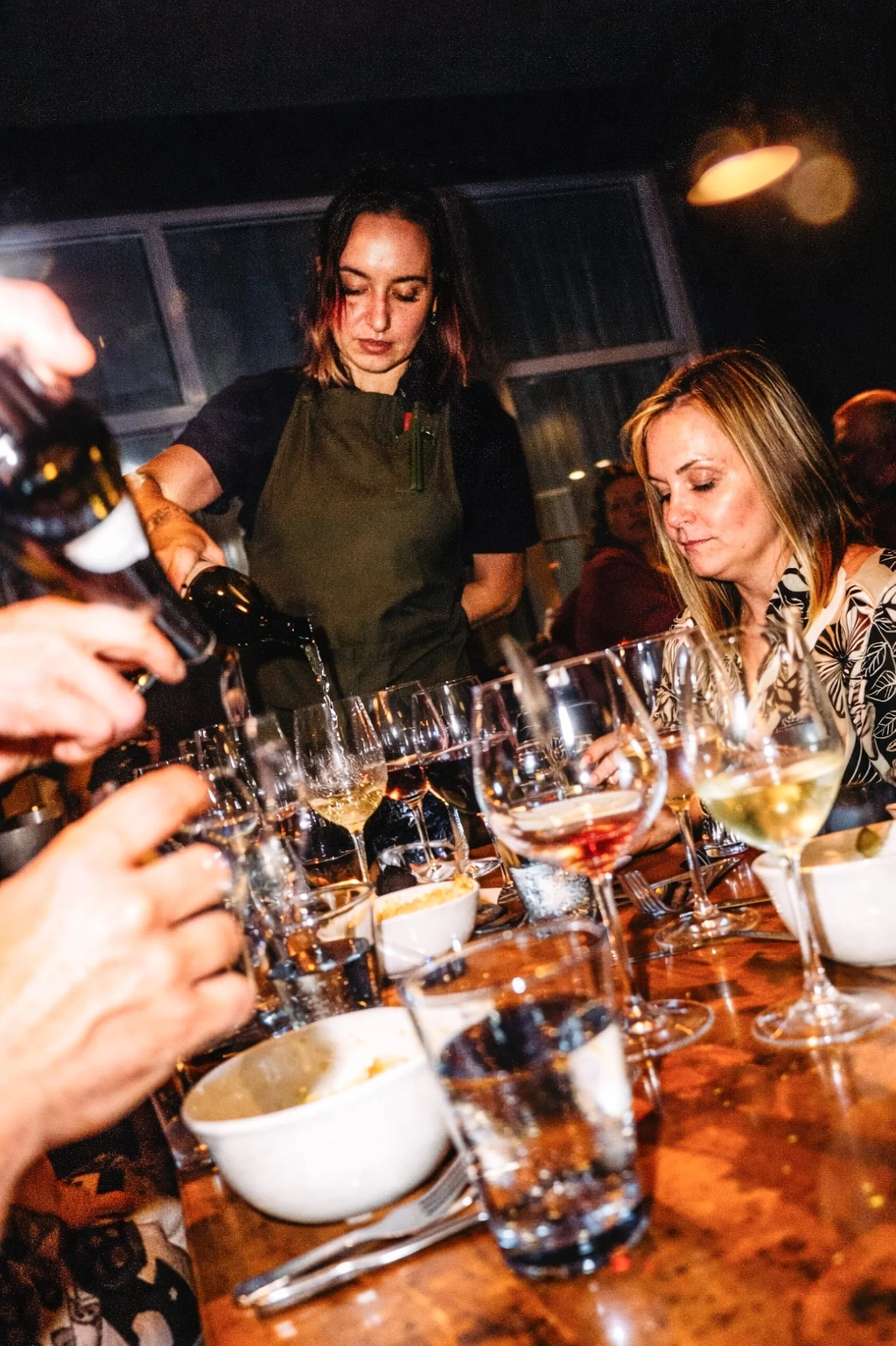 People at a dinner table with wine glasses, bowls of food, and a waitress pouring a drink.