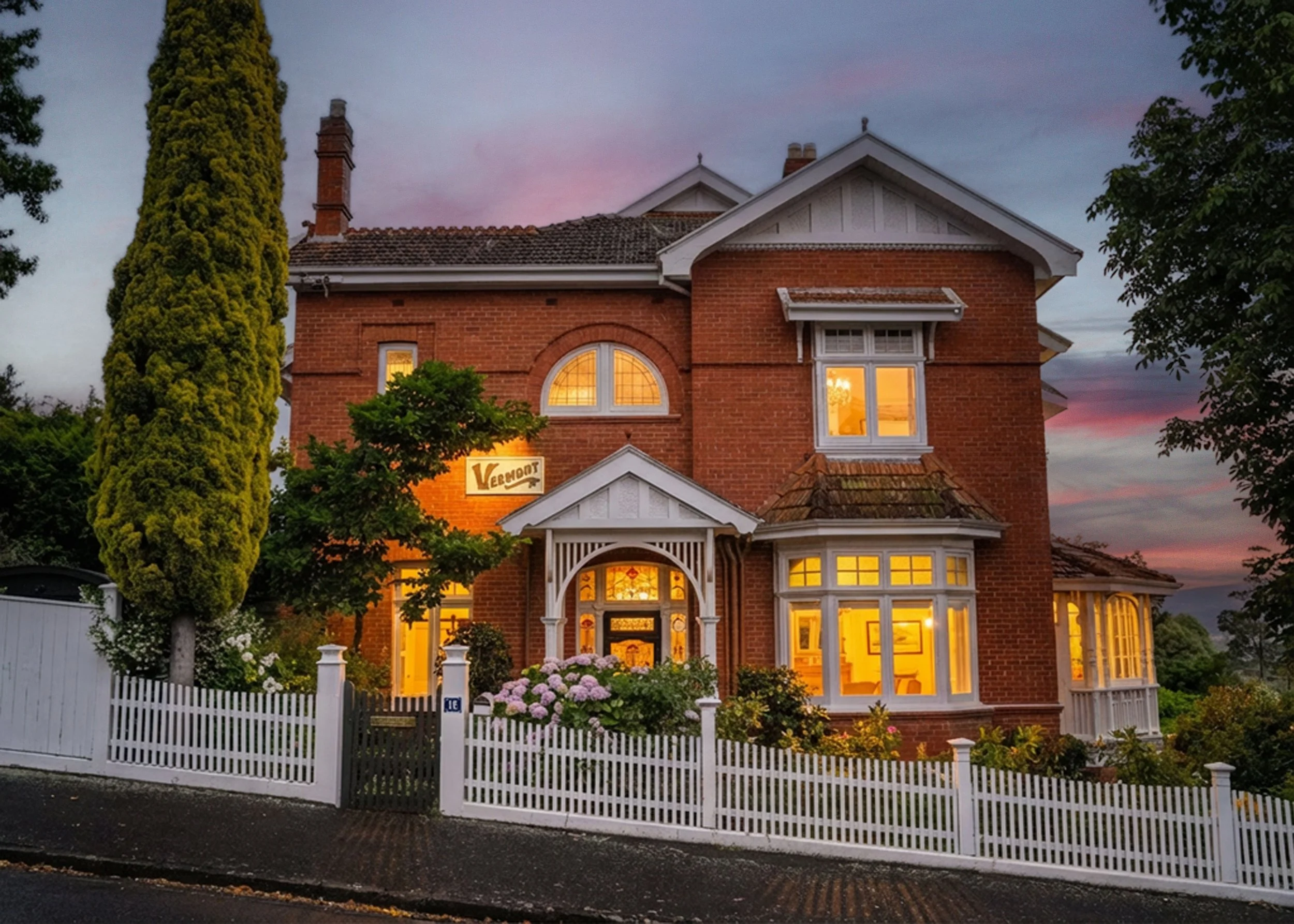 A large brick house with white trim and a white picket fence, illuminated from within, with a purple and pink sunset sky in the background.