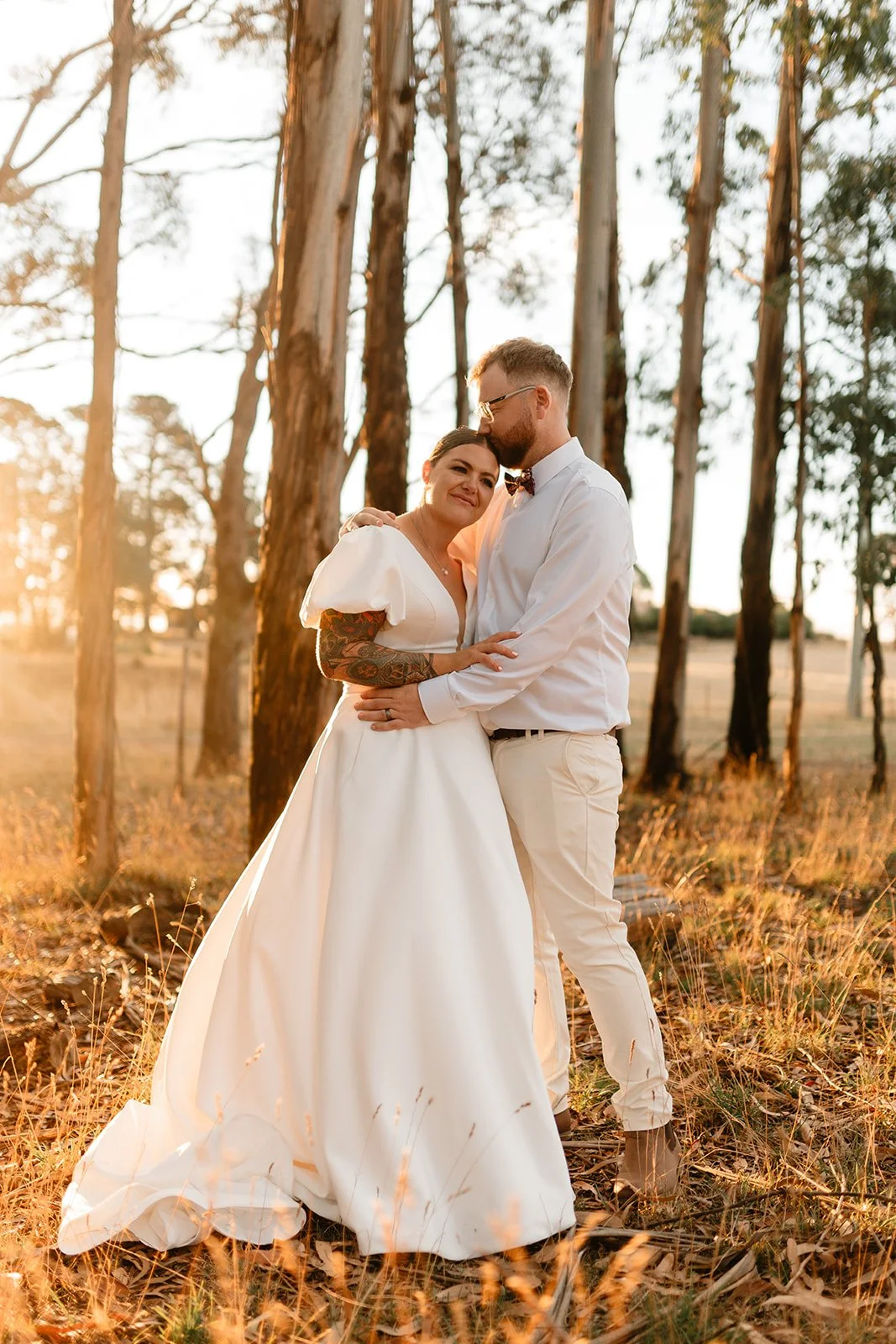 A bride and groom standing close together in a wooded area at sunset, embracing and smiling.