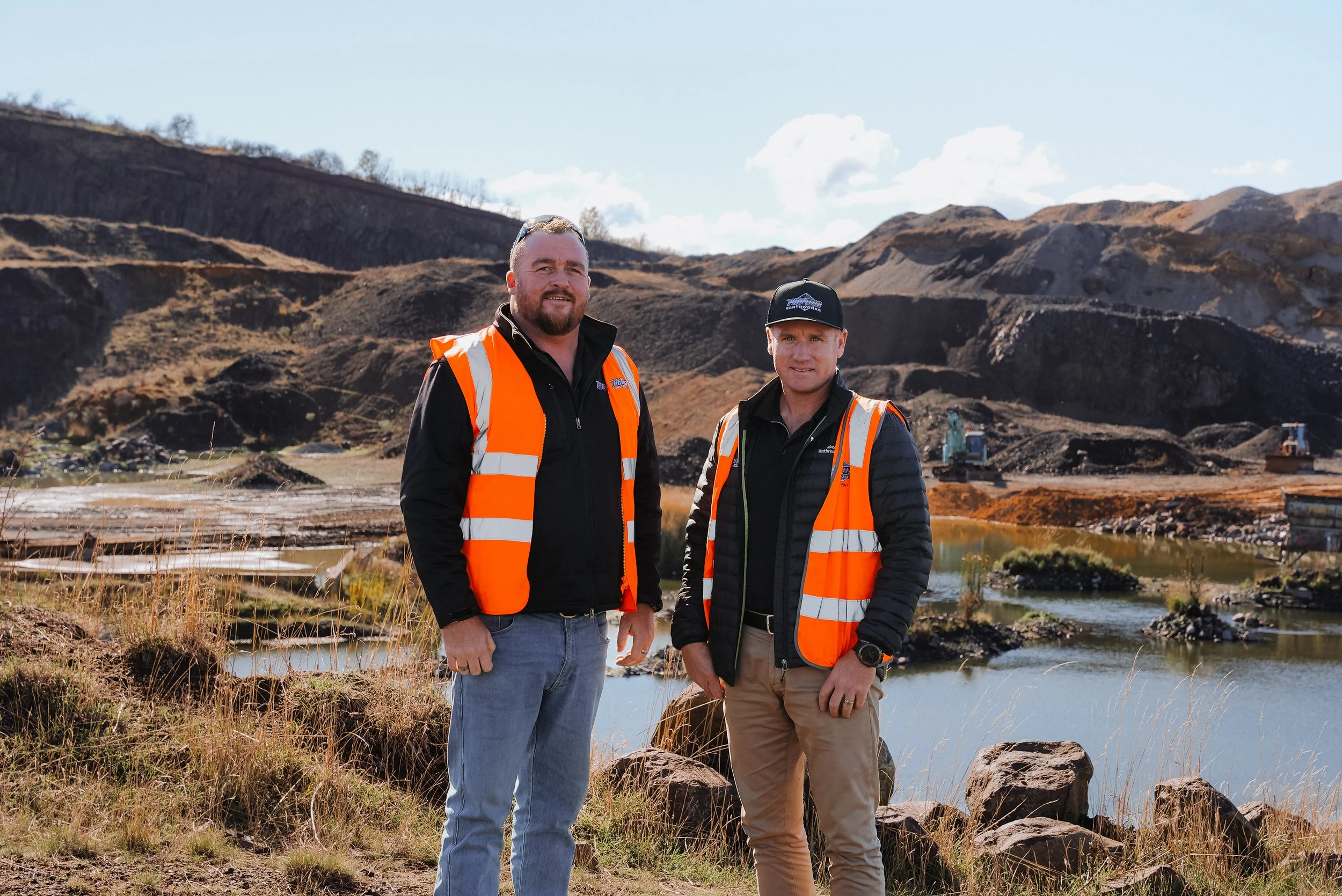 Two men in orange safety vests standing outdoors in a mountainous area with a river and excavation equipment in the background.
