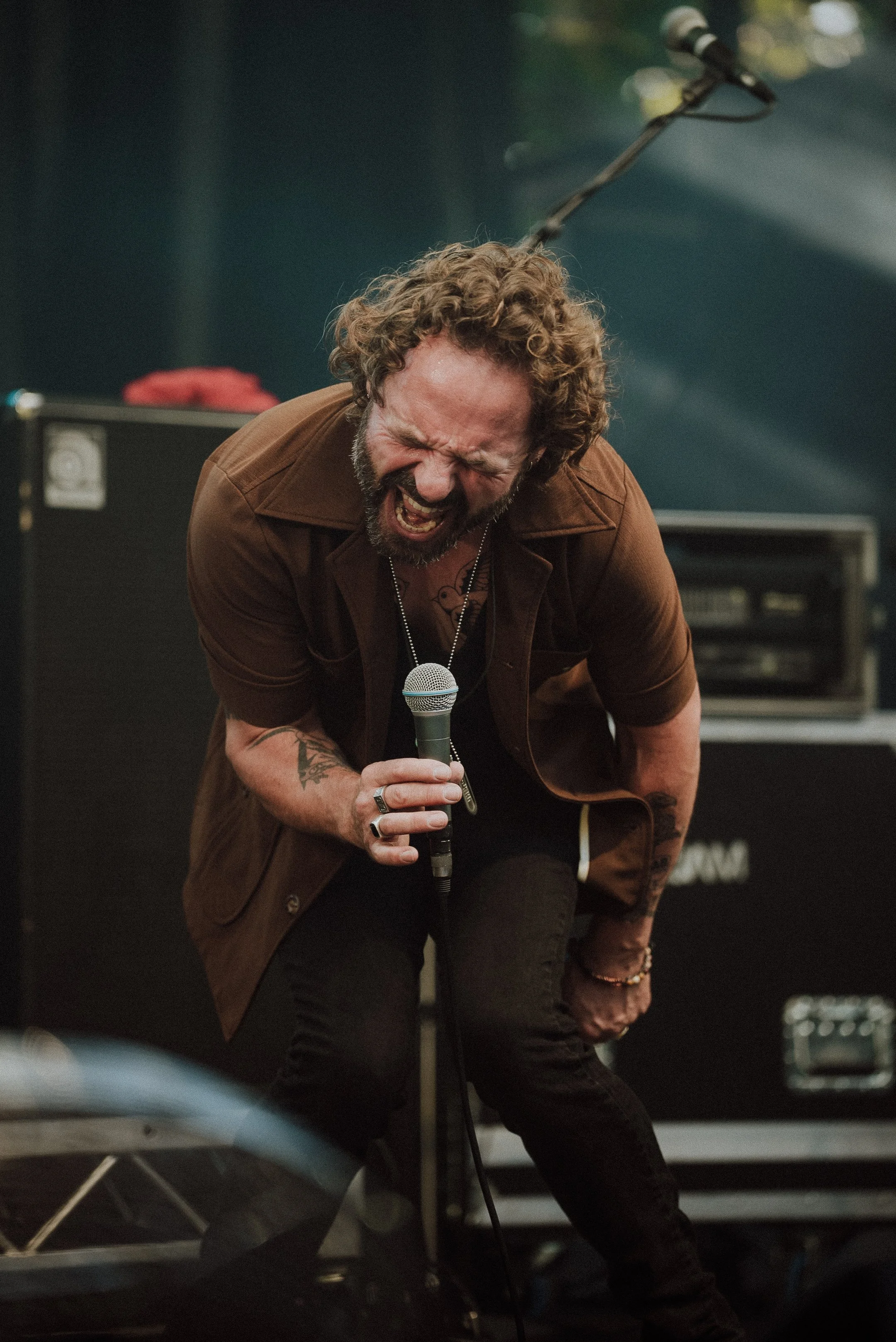 A man with curly hair and a beard passionately singing into a microphone on stage, wearing a brown jacket and black pants.