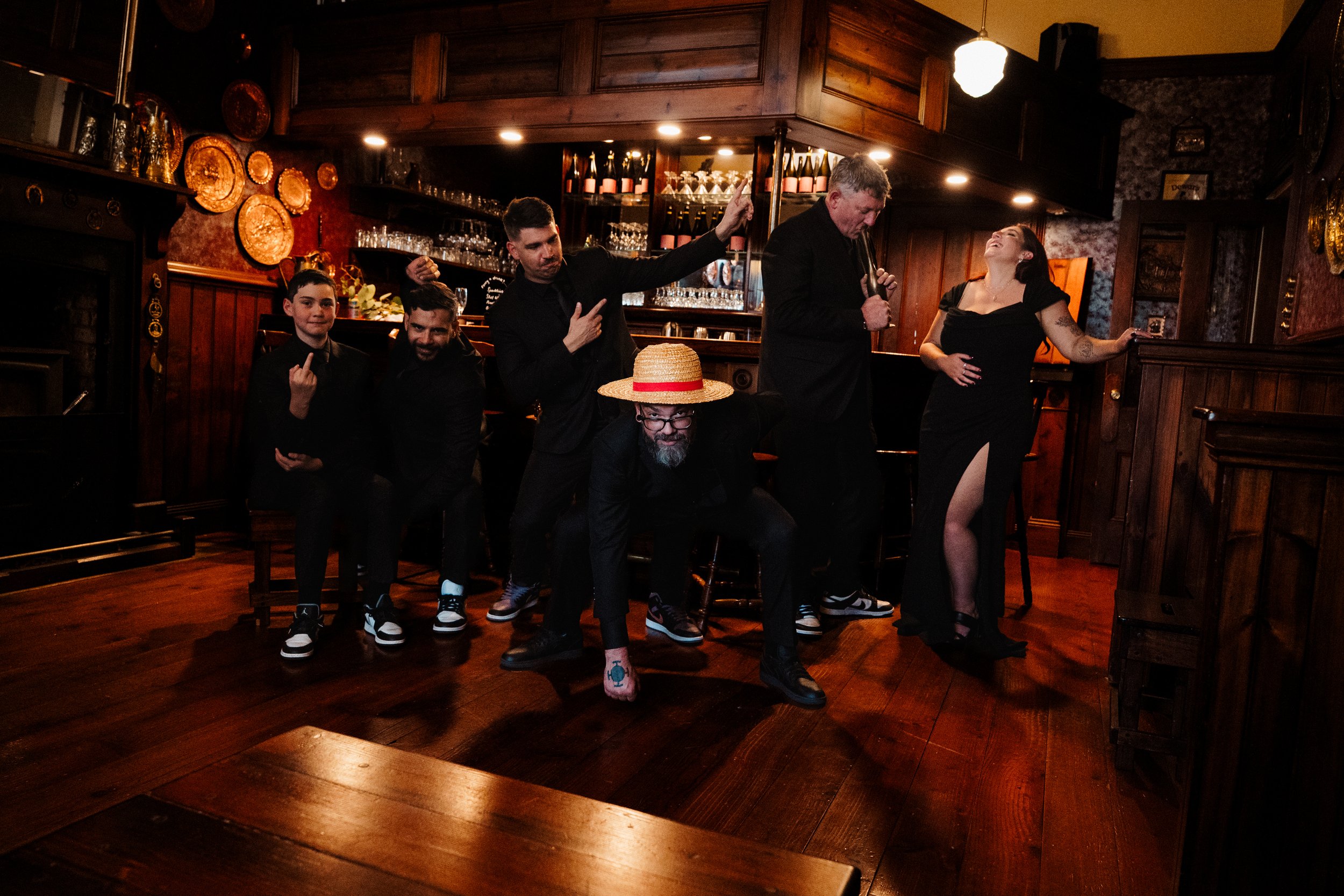 Group of six people dancing and having fun in a dimly lit bar or pub with wooden decor. Four men and two women are dressed in dark clothing, with one man wearing a large straw hat.