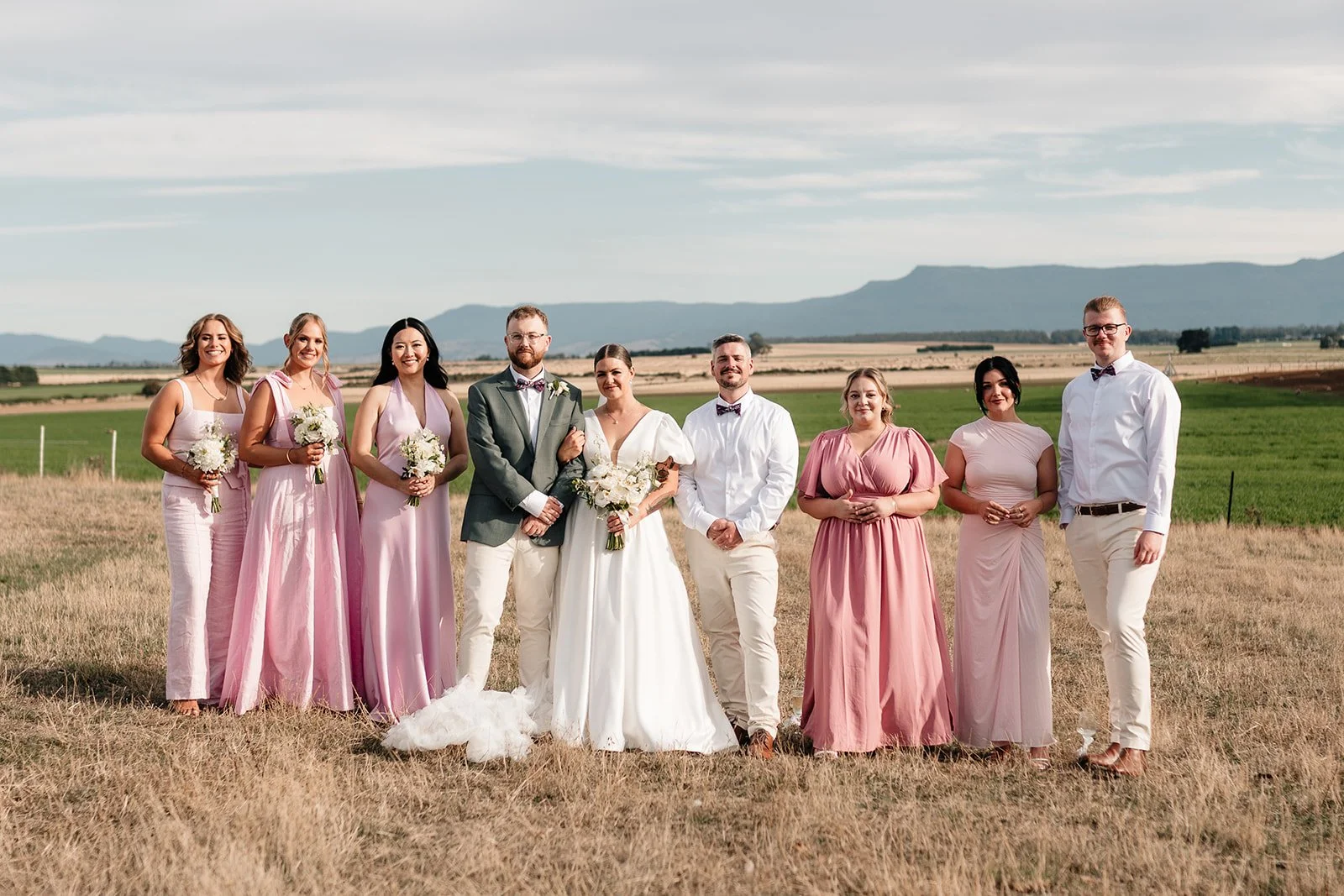 Group of people at a wedding outdoors, dressed in formal attire, with scenic fields and mountains in the background.