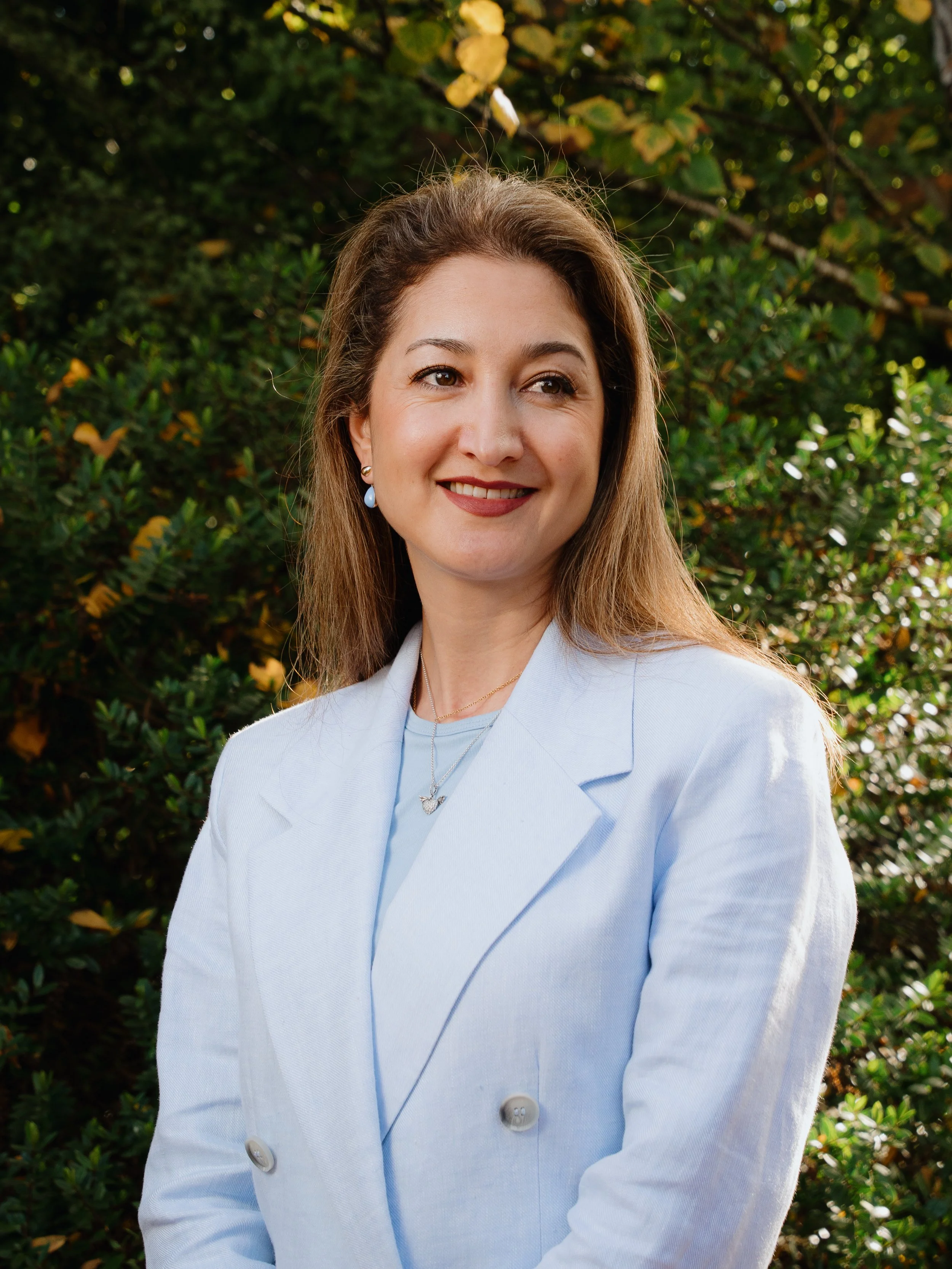 A woman with long brown hair wearing a light blue blazer, standing outdoors in front of green foliage, smiling and looking to her right.