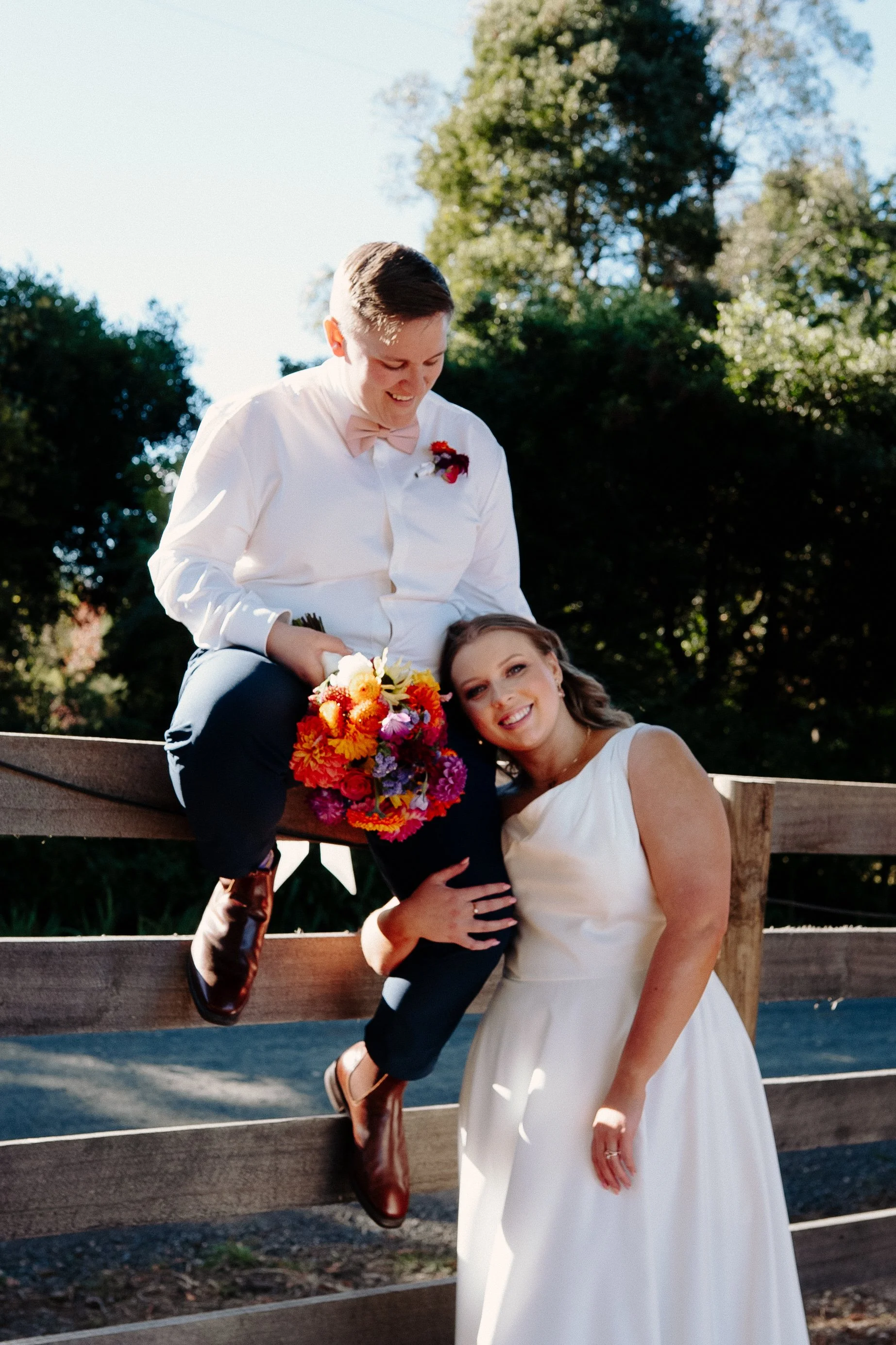 A wedding couple poses outdoors near a wooden fence, with a guy sitting on the fence holding a colorful bouquet and a woman standing beside him, smiling, in a white dress. The background includes tall trees and a clear sky.