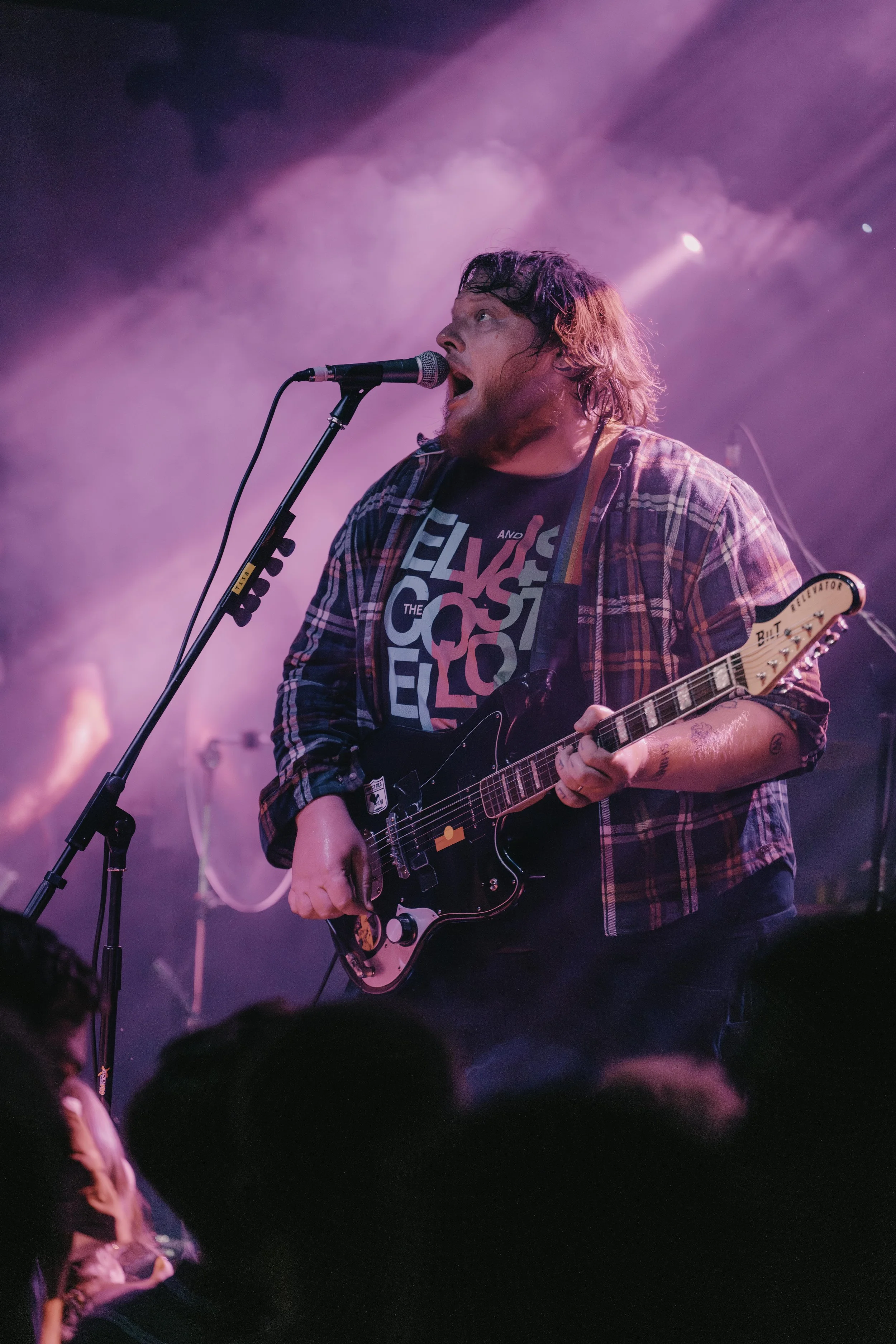 Male musician performing on stage with a guitar and microphone, wearing a plaid shirt and graphic T-shirt, purple and pink stage lighting.