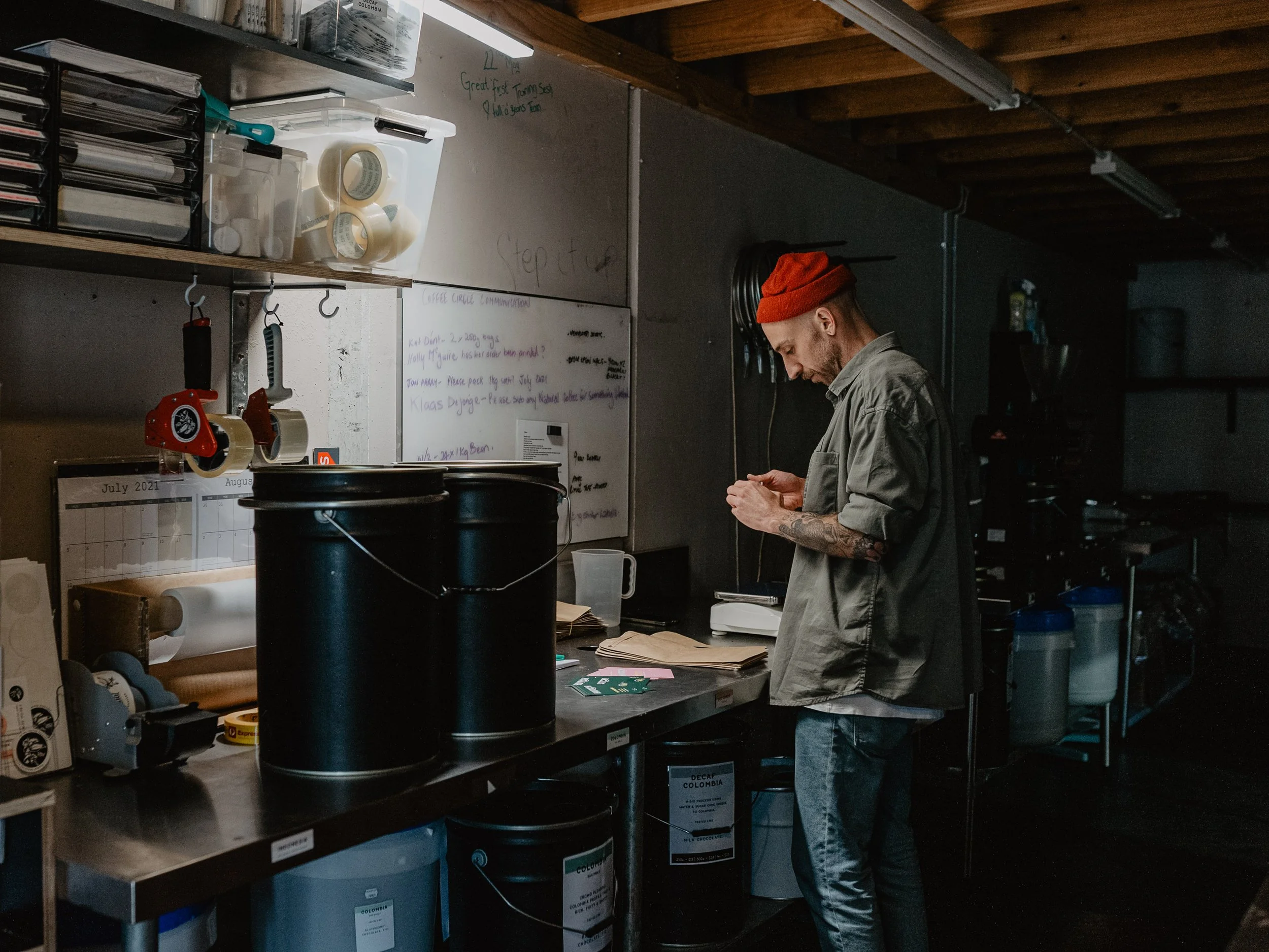 A man with a red beanie, gray shirt, and tattoos on his left forearm is standing at a workbench in a workshop or kitchen, looking down while working on something with his hands. The background shows shelves with containers, a whiteboard with handwrit
